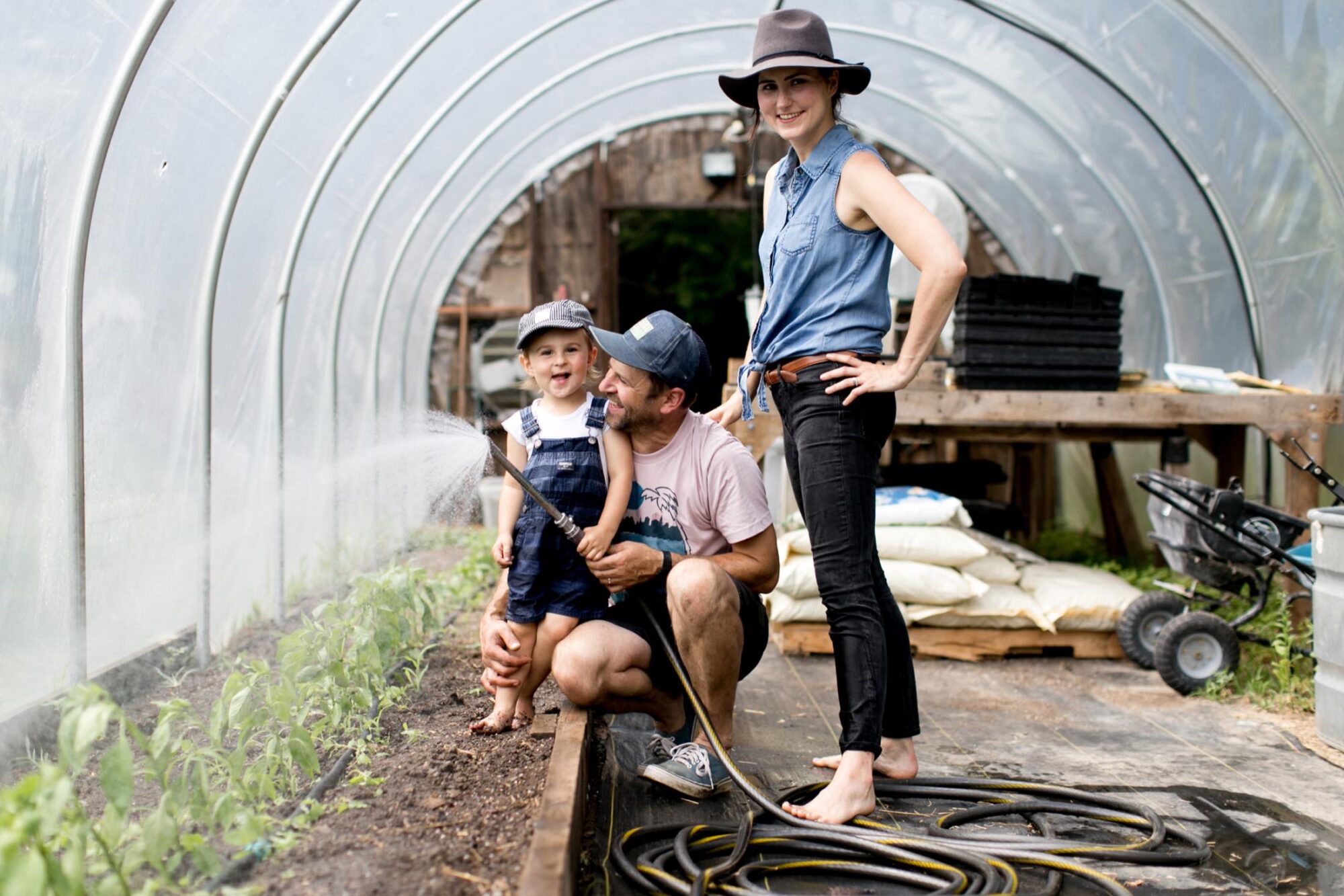 Seattle's best commercial food and hospitality photographer A family of three inside a greenhouse waters young plants. Captured by Seattles best commercial food lifestyle photographer, the father holds a toddler, both smiling, while the mother stands nearby, hands on hips. Gardening tools and supplies are visible in the background. captured by seattle's best food and lifestyle photographer Brooke Fitts