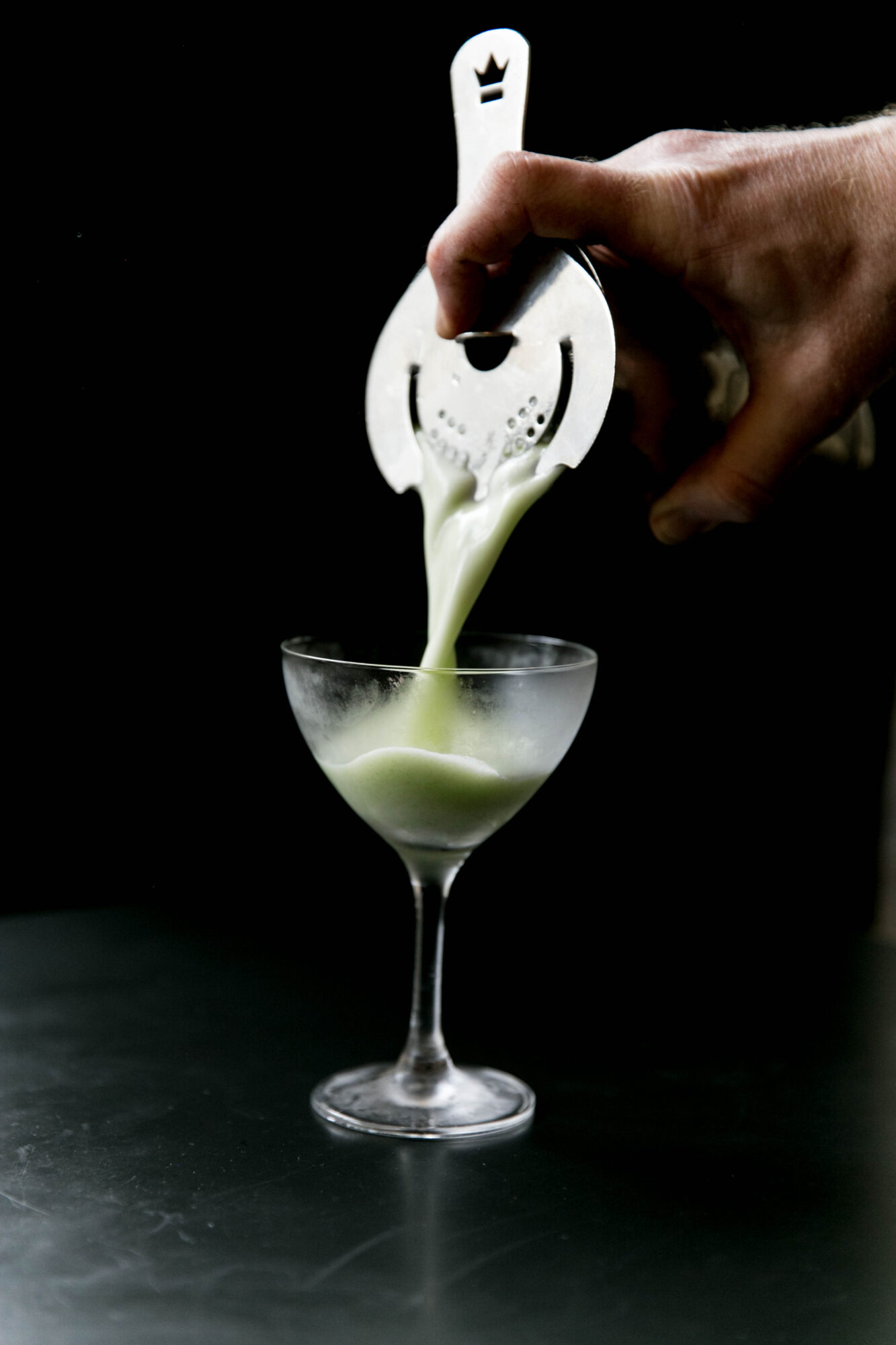 Seattle's best commercial food and hospitality photographer A hand pours a pale green cocktail through a strainer into a coupe glass, against a dark background—captured by Seattles best commercial food lifestyle photographer. captured by seattle's best food and lifestyle photographer Brooke Fitts