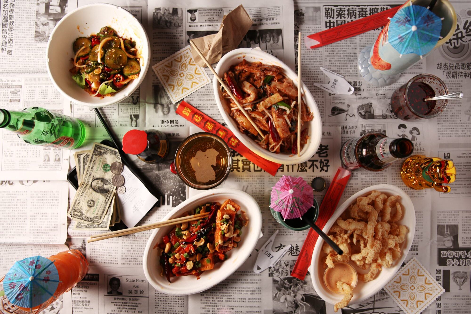 Seattle's best commercial food and hospitality photographer A table covered with Chinese newspapers holds various Asian dishes, drinks with umbrellas, chopsticks, soy sauce, U.S. dollar bills, and soda bottles—captured by Seattles best commercial food lifestyle photographer for a vibrant dining scene from above. captured by seattle's best food and lifestyle photographer Brooke Fitts
