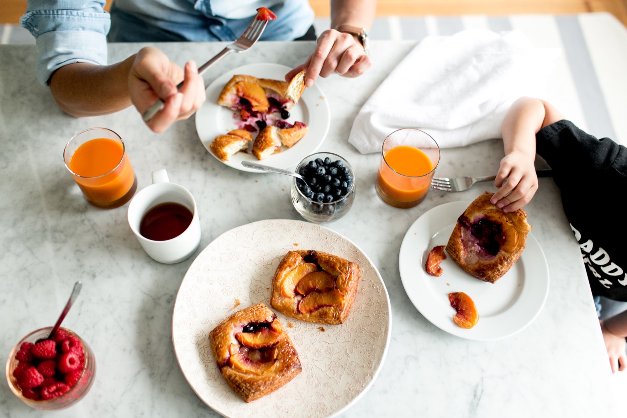 Seattle's best commercial food and hospitality photographer A breakfast scene by Seattles best commercial food lifestyle photographer: two people enjoy fruit pastries at a marble table, with orange juice, tea, fresh raspberries, blueberries, and a white napkin. captured by seattle's best food and lifestyle photographer Brooke Fitts