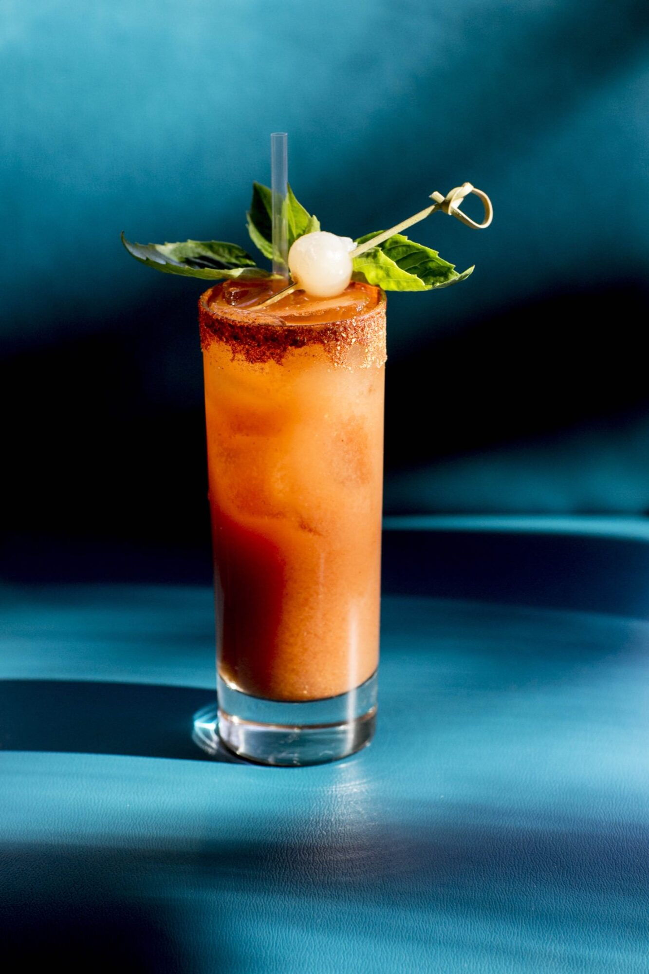 Seattle's best commercial food and hospitality photographer Captured by Seattles best commercial food lifestyle photographer, a tall glass of orange-red cocktail with ice, garnished with green leaves and a white cherry on a skewer, sits on a blue surface with a matching blue background. captured by seattle's best food and lifestyle photographer Brooke Fitts