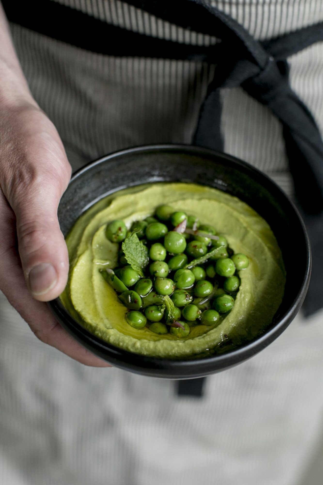 Seattle's best commercial food and hospitality photographer A person wearing a striped apron holds a black bowl filled with green hummus topped with fresh green peas, mint leaves, and a drizzle of olive oil, captured by Seattles best commercial food lifestyle photographer. captured by seattle's best food and lifestyle photographer Brooke Fitts
