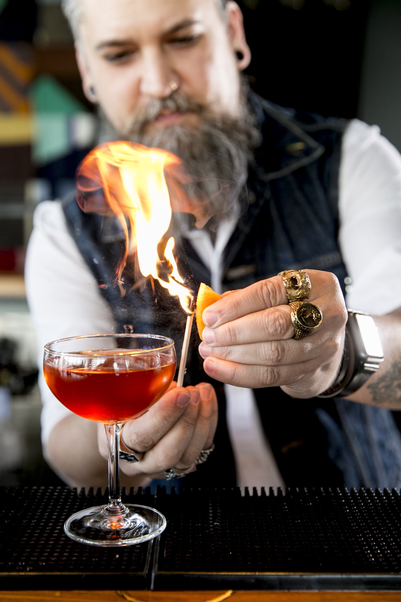 Seattle's best commercial food and hospitality photographer A bartender with a beard and rings on his fingers, captured by Seattles best commercial food lifestyle photographer, lights an orange peel with a match over a cocktail, creating a burst of flame above the drink. captured by seattle's best food and lifestyle photographer Brooke Fitts