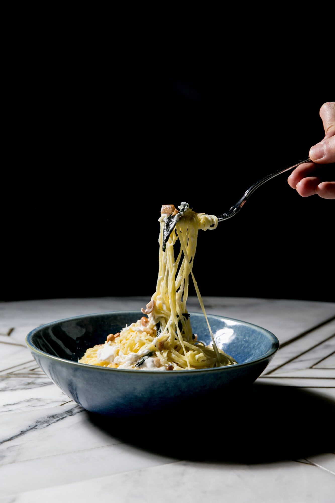 Seattle's best commercial food and hospitality photographer A hand lifts a forkful of creamy pasta from a blue bowl on a marble table, captured by Seattles best commercial food lifestyle photographer against a dark background. captured by seattle's best food and lifestyle photographer Brooke Fitts