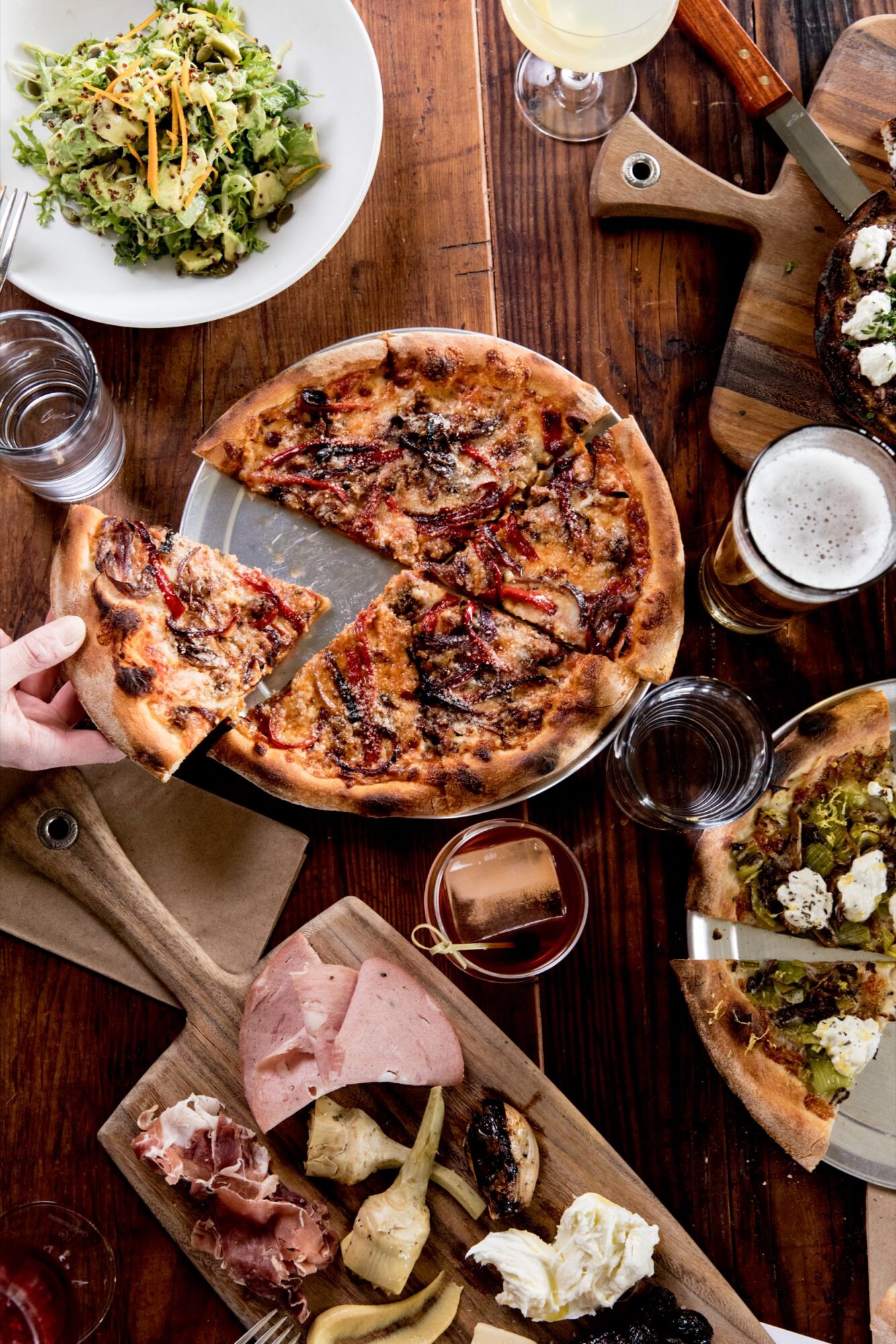 Seattle's best commercial food and hospitality photographer A table set with a pizza being served, a green salad, charcuterie board, another pizza, drinks, and utensils on a wooden surface—captured by Seattles best commercial food lifestyle photographer. captured by seattle's best food and lifestyle photographer Brooke Fitts
