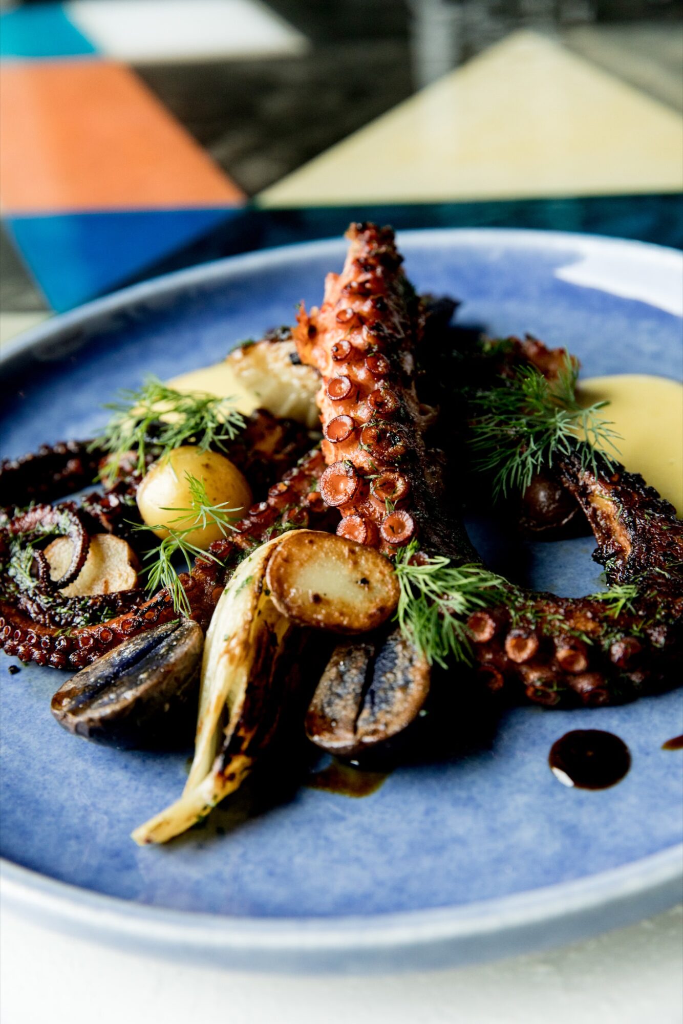 Seattle's best commercial food and hospitality photographer Grilled octopus tentacles served on a blue plate with roasted vegetables, fresh herbs, and a light sauce, beautifully captured by Seattles best commercial food lifestyle photographer in a stylish, modern setting. captured by seattle's best food and lifestyle photographer Brooke Fitts