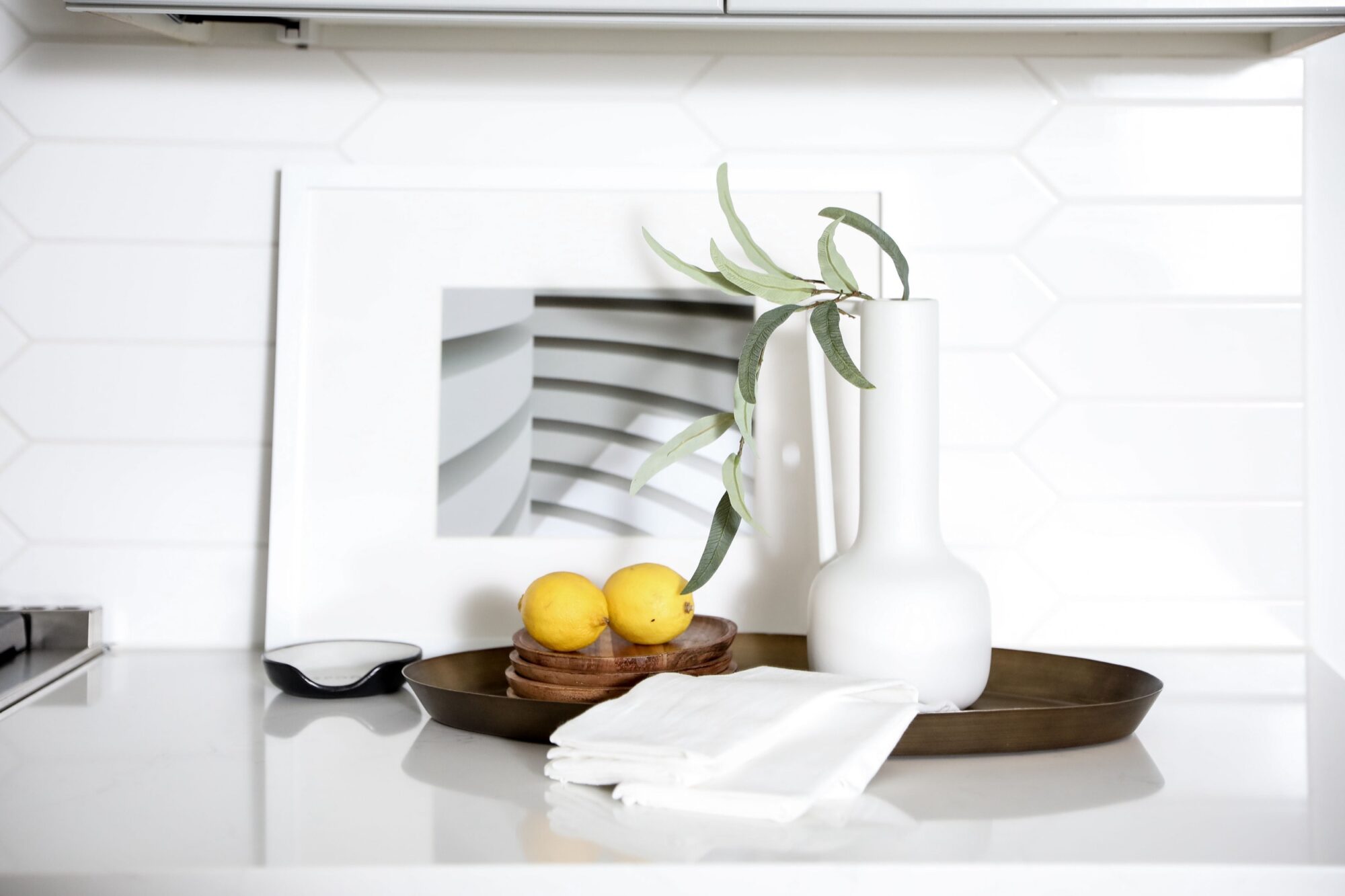 Seattle's best commercial food and hospitality photographer A modern white kitchen counter styled by Seattles best commercial food lifestyle photographer features a white vase with green leaves, two lemons, stacked wooden coasters, a folded towel, and abstract art against a white tile backsplash. captured by seattle's best food and lifestyle photographer Brooke Fitts