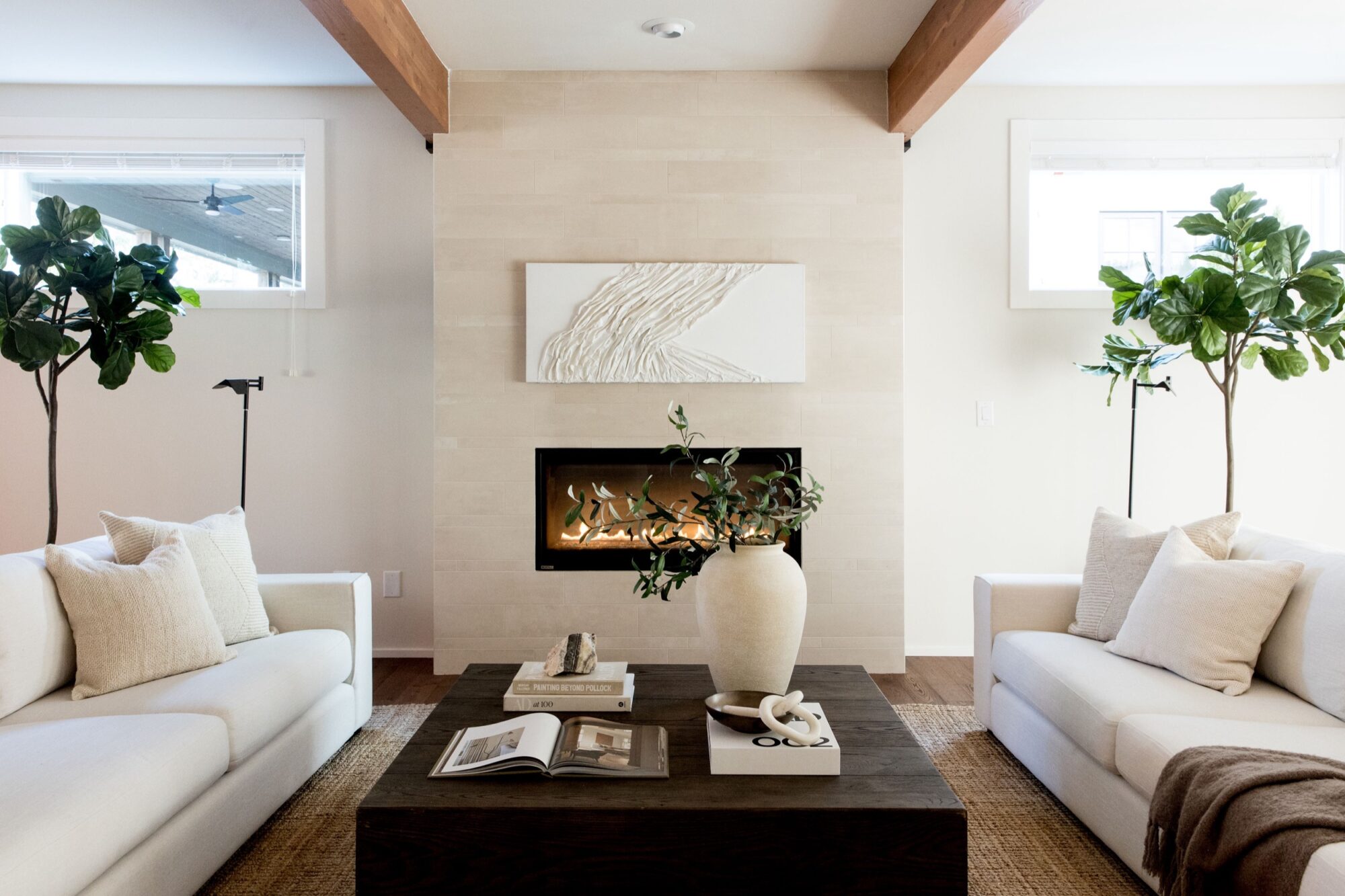 Seattle's best commercial food and hospitality photographer A modern living room with two white sofas, a dark wooden coffee table with books and a vase, a fireplace with abstract art above it, and large potted plants—captured by Seattles best commercial food lifestyle photographer—in soft neutral tones. captured by seattle's best food and lifestyle photographer Brooke Fitts