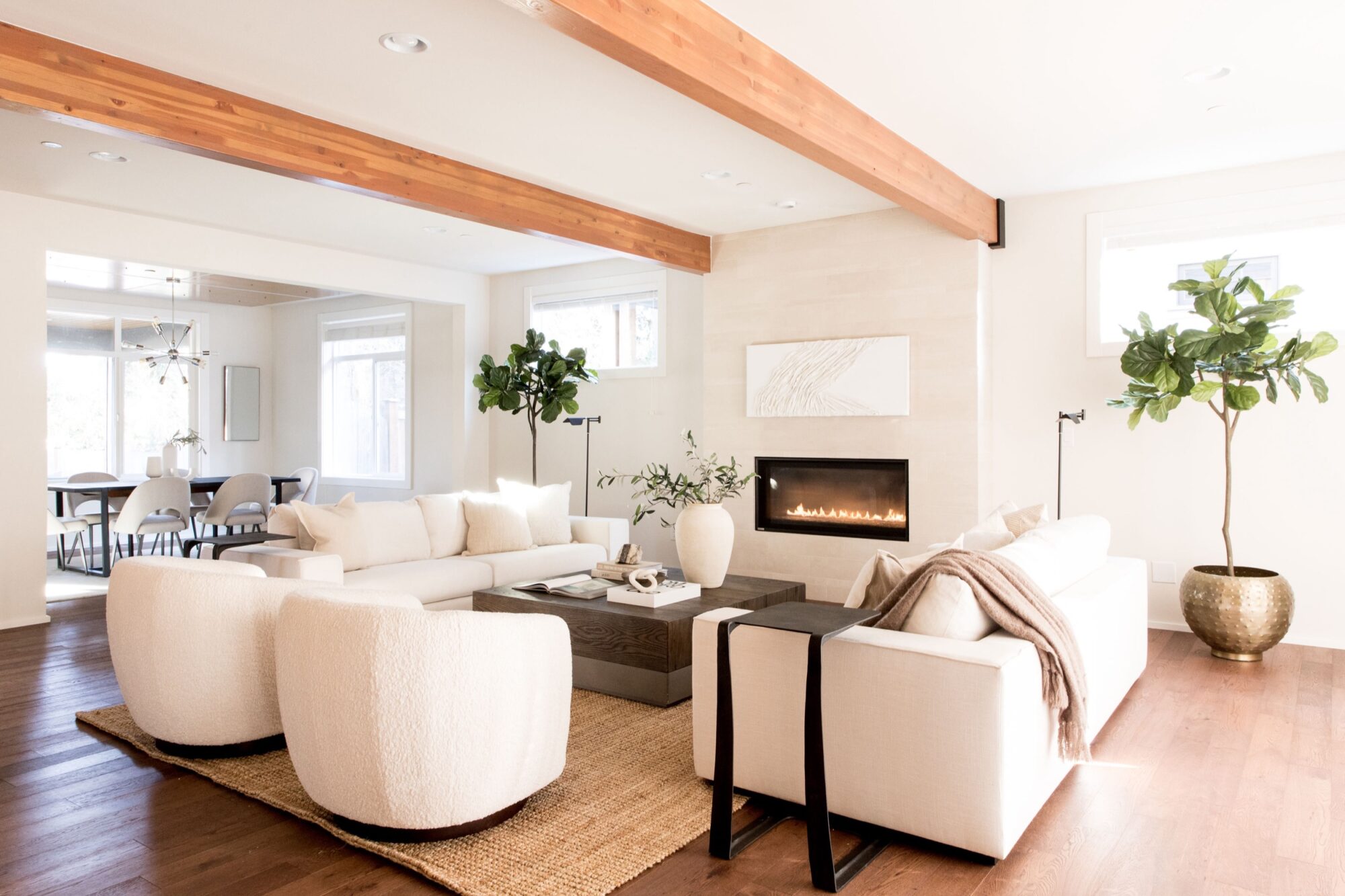 Seattle's best commercial food and hospitality photographer A modern living room with white sofas and armchairs, a beige rug, fireplace, large potted plants, wooden ceiling beams, and abundant natural light, captured by Seattles best commercial food lifestyle photographer; a dining area is visible in the background. captured by seattle's best food and lifestyle photographer Brooke Fitts