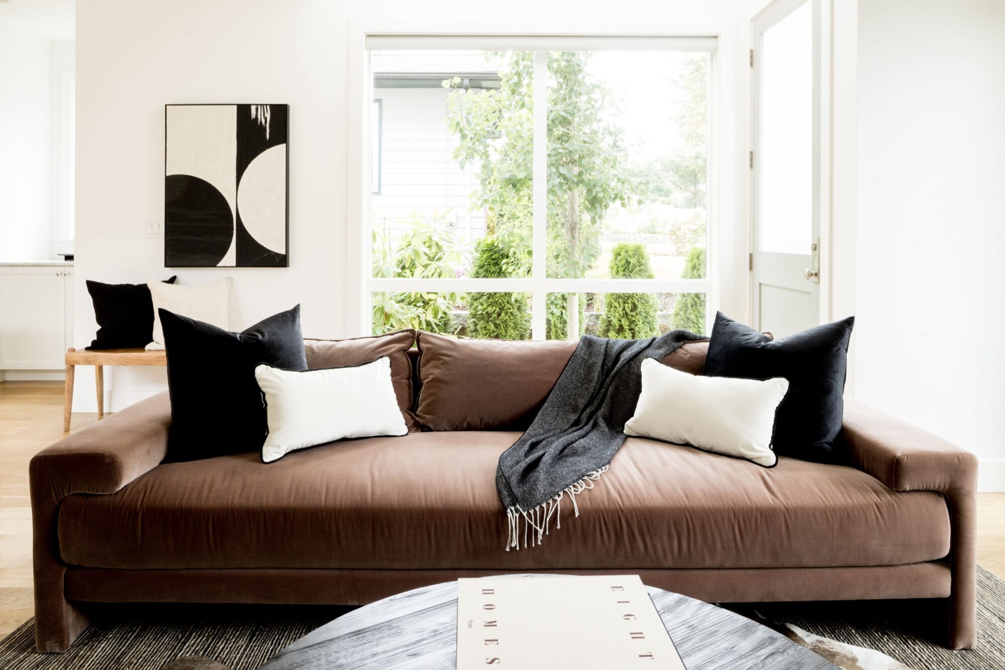 Seattle's best commercial food and hospitality photographer A modern living room with a brown sofa decorated with black and white pillows, a dark throw blanket, and minimalist wall art, captured by Seattles best commercial food lifestyle photographer. Large windows let in natural light, with a door open to a green outdoor area. captured by seattle's best food and lifestyle photographer Brooke Fitts