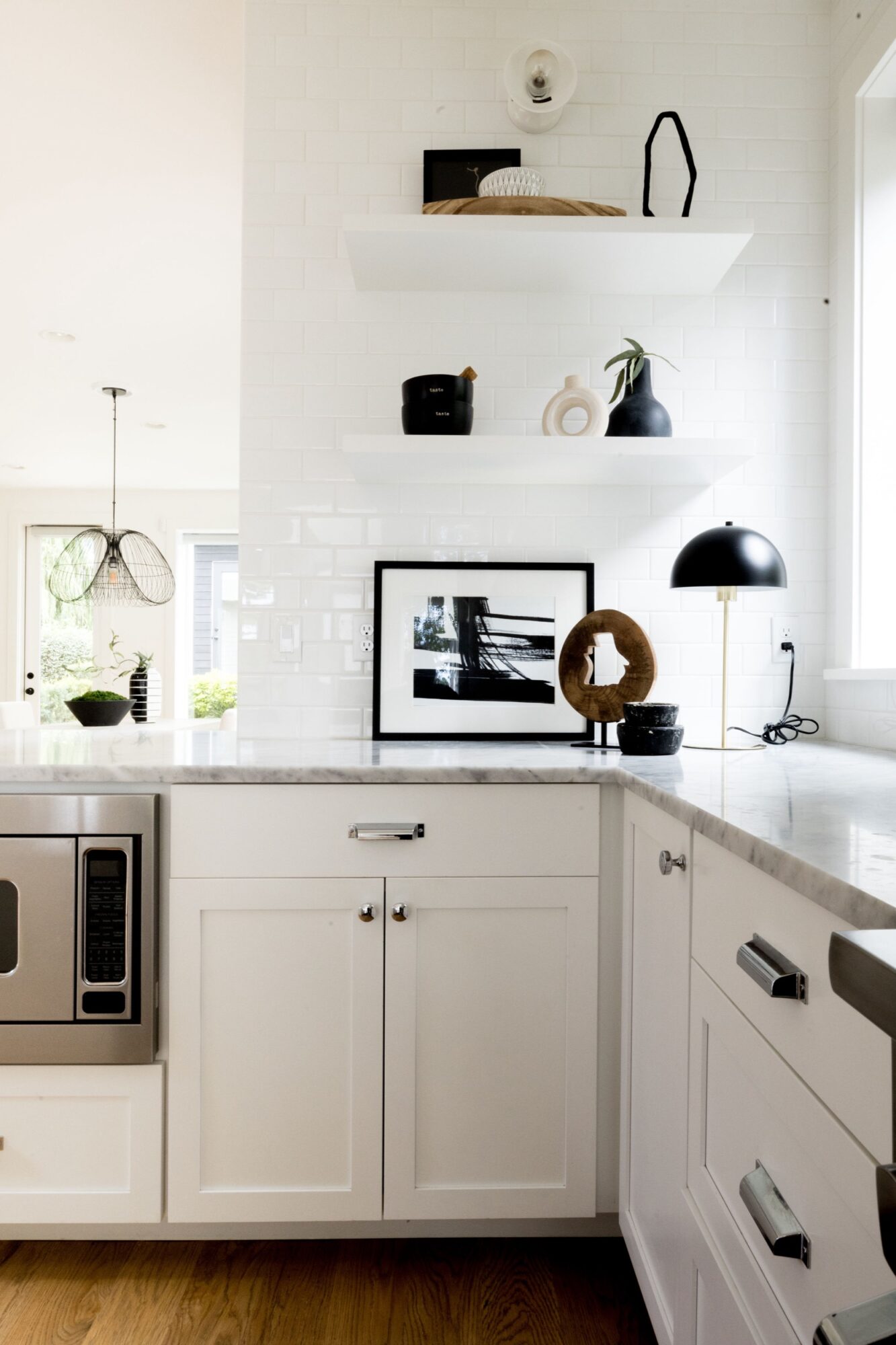 Seattle's best commercial food and hospitality photographer Modern white kitchen with sleek cabinets, marble countertop, wooden floor, and stylish decor—captured by Seattles best commercial food lifestyle photographer. Natural light highlights the elegant shelves, framed photo, black table lamp, and built-in microwave. captured by seattle's best food and lifestyle photographer Brooke Fitts