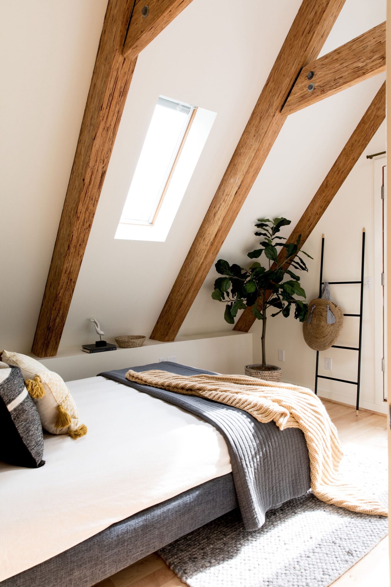 Seattle's best commercial food and hospitality photographer A cozy attic bedroom with exposed wooden beams, a skylight, a gray bed with neutral-toned pillows and blankets, a potted plant, and a black ladder shelf—captured by Seattles best commercial food lifestyle photographer. captured by seattle's best food and lifestyle photographer Brooke Fitts