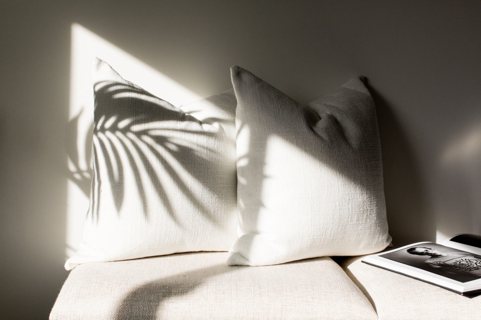 Seattle's best commercial food and hospitality photographer Two white textured pillows on a beige seat with sunlight casting a leafy shadow—styled by Seattles best commercial food lifestyle photographer. An open book featuring a black-and-white photo lies at the corner, capturing cozy elegance. captured by seattle's best food and lifestyle photographer Brooke Fitts