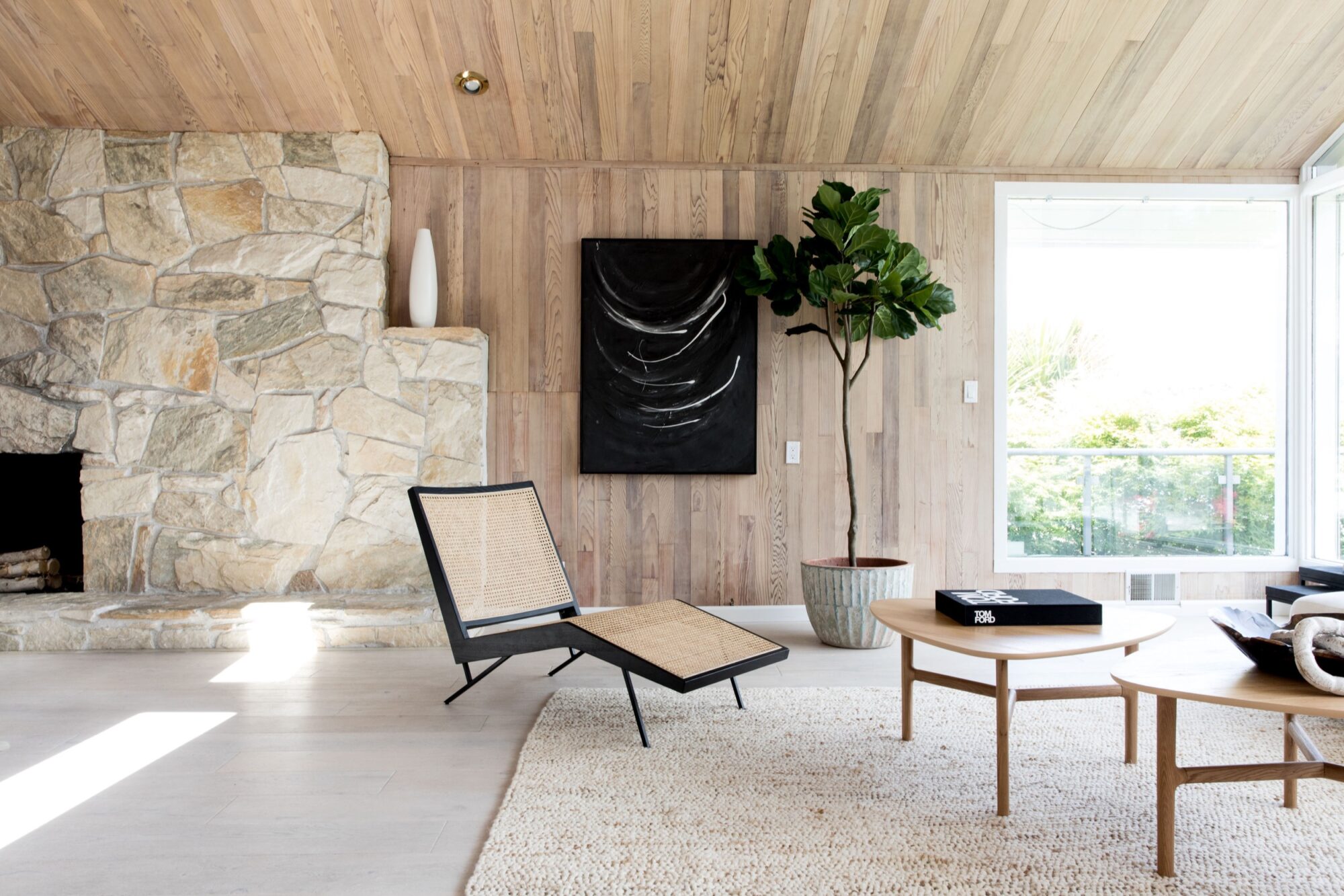 Seattle's best commercial food and hospitality photographer A modern living room with light wood walls and ceiling, a stone fireplace, black lounge chair with woven seat, large potted plant, round wooden tables, and a black abstract painting—captured by Seattles best commercial food lifestyle photographer. captured by seattle's best food and lifestyle photographer Brooke Fitts