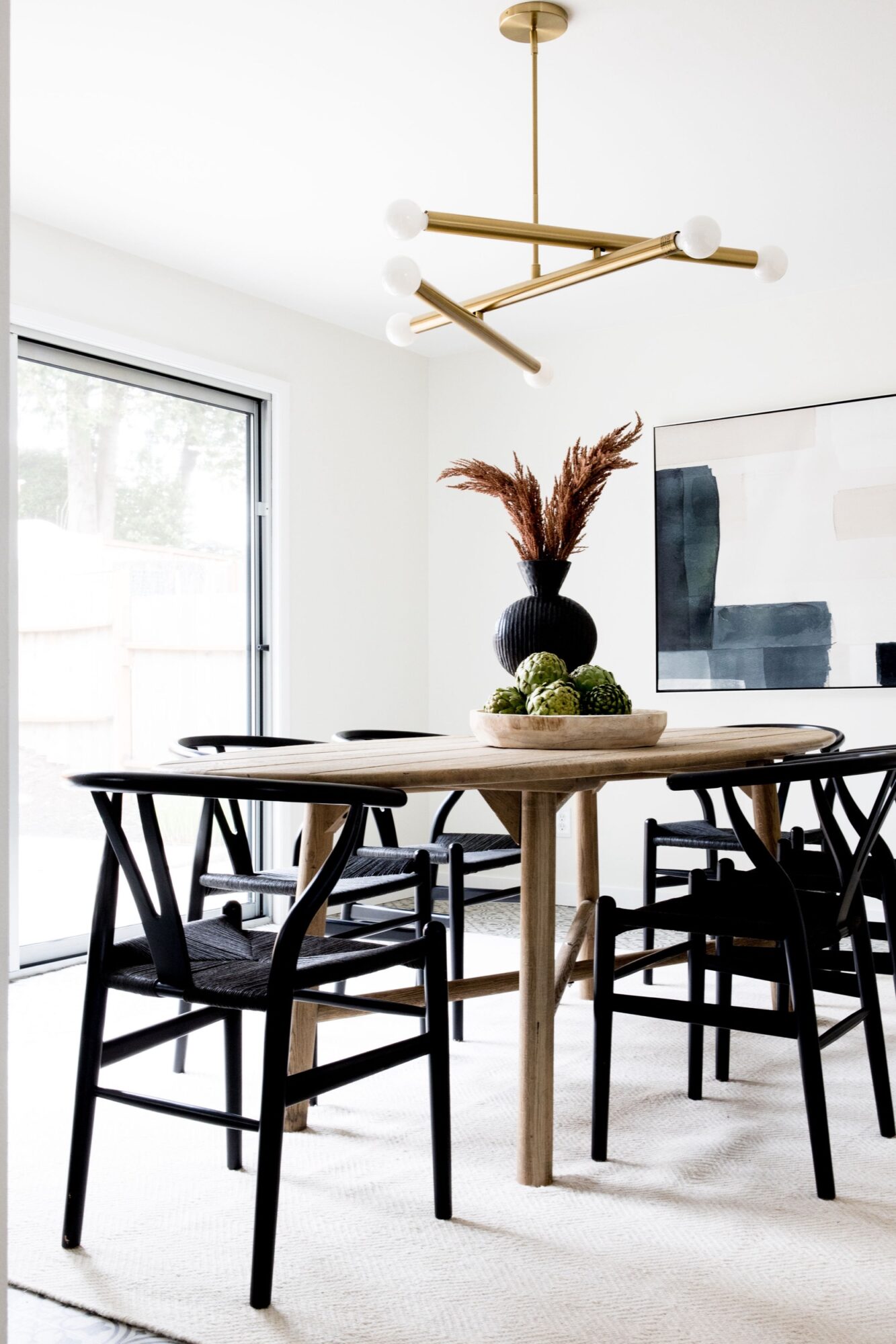 Seattle's best commercial food and hospitality photographer A modern dining room with a round wooden table, black chairs, a black vase with dried plants, artichokes on a tray, abstract wall art, large window, and gold geometric chandelier—captured by Seattles best commercial food lifestyle photographer. captured by seattle's best food and lifestyle photographer Brooke Fitts