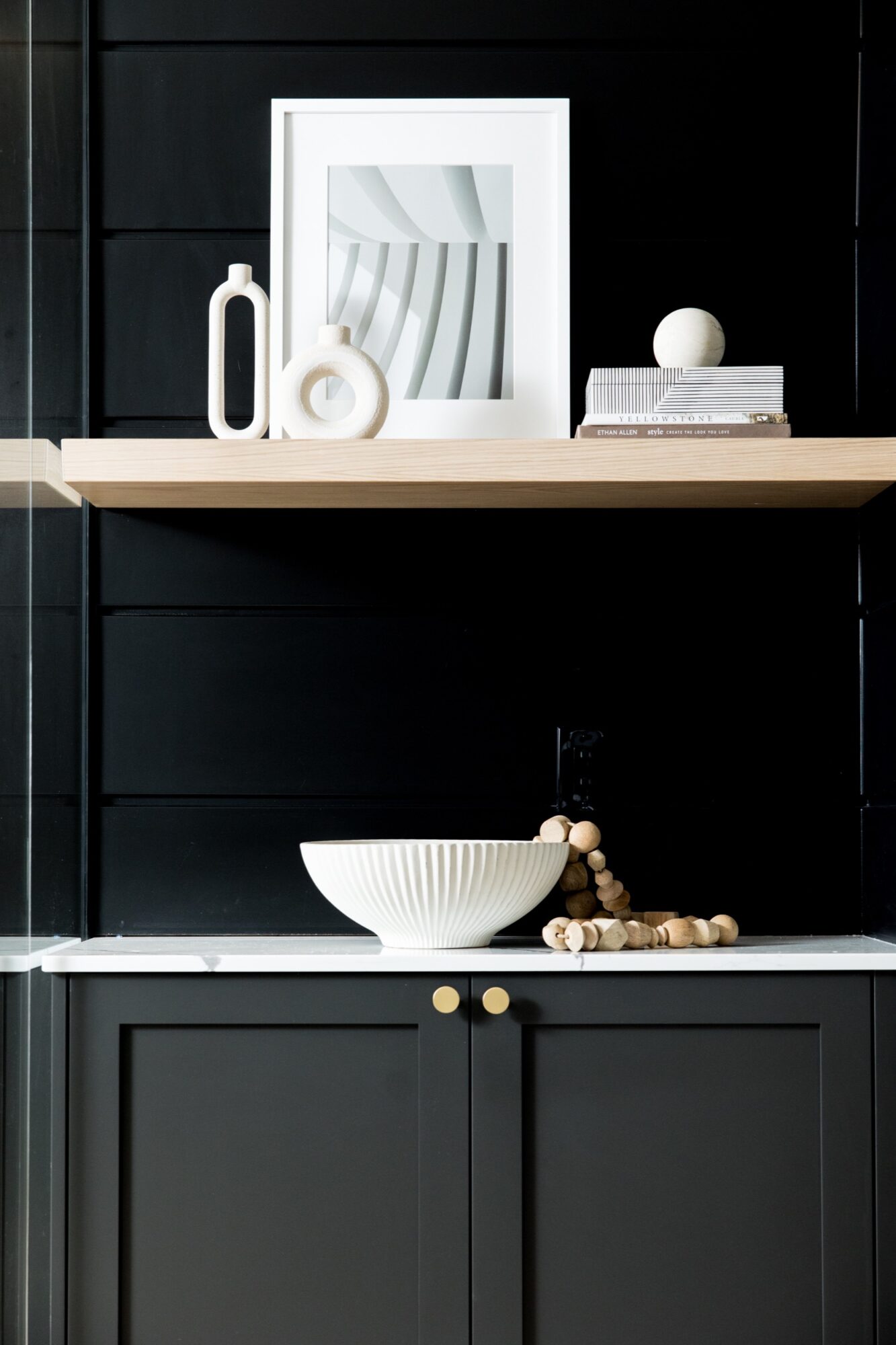 Seattle's best commercial food and hospitality photographer A modern bathroom with black cabinets, a white ribbed vessel sink, and wooden beads on the counter—captured by Seattles best commercial food lifestyle photographer. Above, a light wood shelf displays art, books, and unique decor. captured by seattle's best food and lifestyle photographer Brooke Fitts