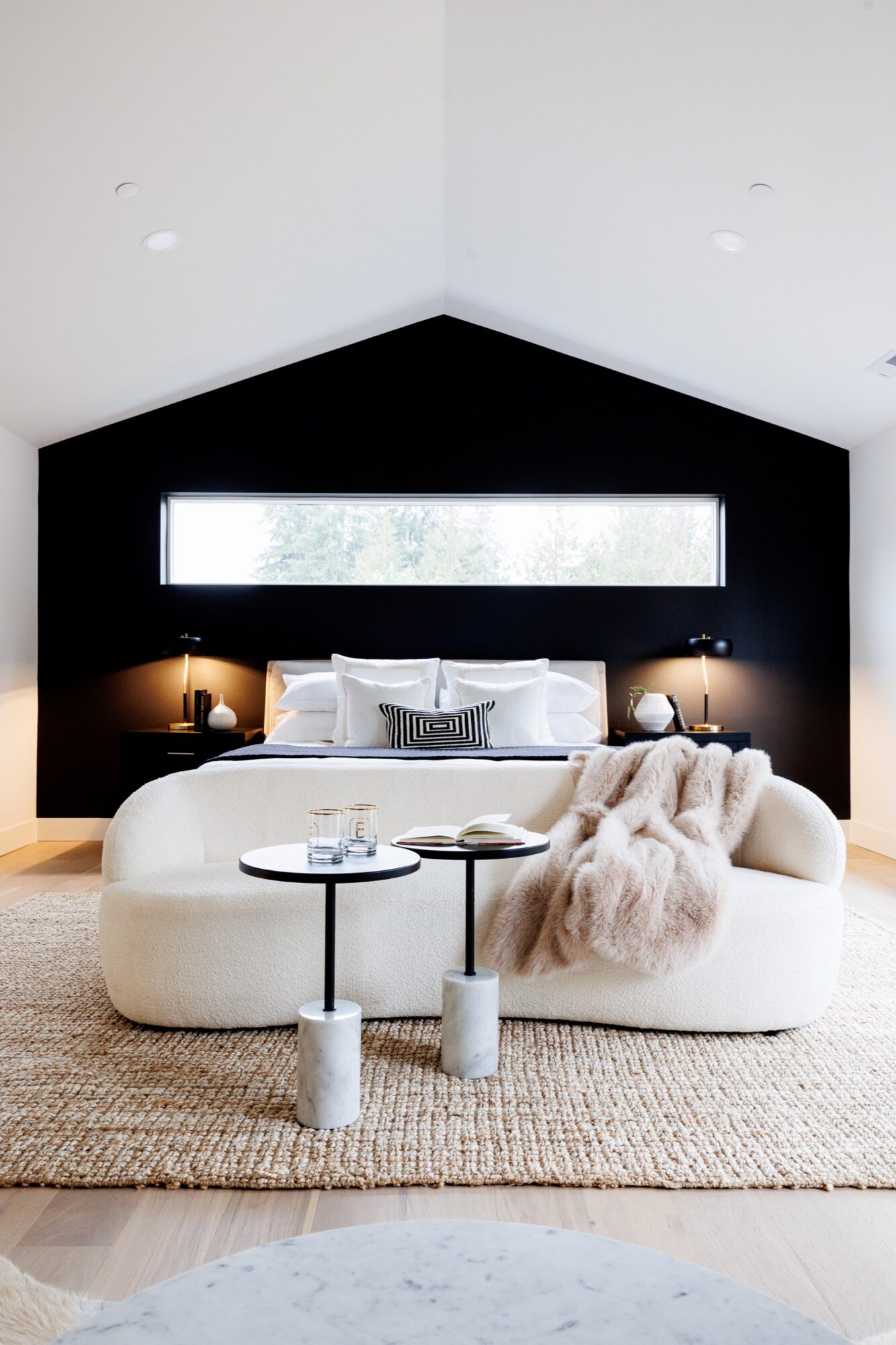 Seattle's best commercial food and hospitality photographer A modern bedroom with a black accent wall, white bedding, and a cream-colored curved sofa facing the bed—styled like Seattles best commercial food lifestyle photographer would capture it. Natural light streams in from a large window onto a cozy beige rug. captured by seattle's best food and lifestyle photographer Brooke Fitts