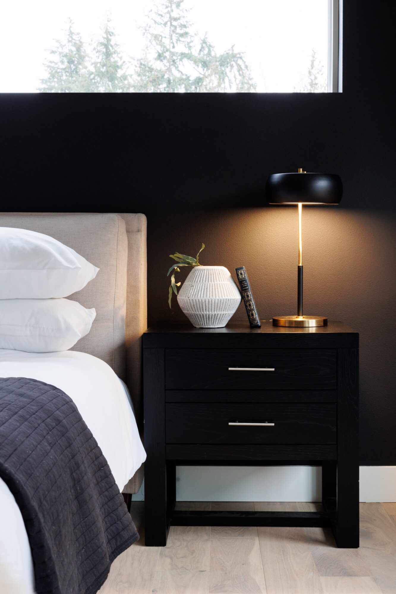 Seattle's best commercial food and hospitality photographer A modern bedroom nightstand with two drawers, styled by Seattles best commercial food lifestyle photographer, holds a white textured vase with a plant, a book, and a sleek black and gold lamp beside a bed with white bedding and a grey headboard. captured by seattle's best food and lifestyle photographer Brooke Fitts