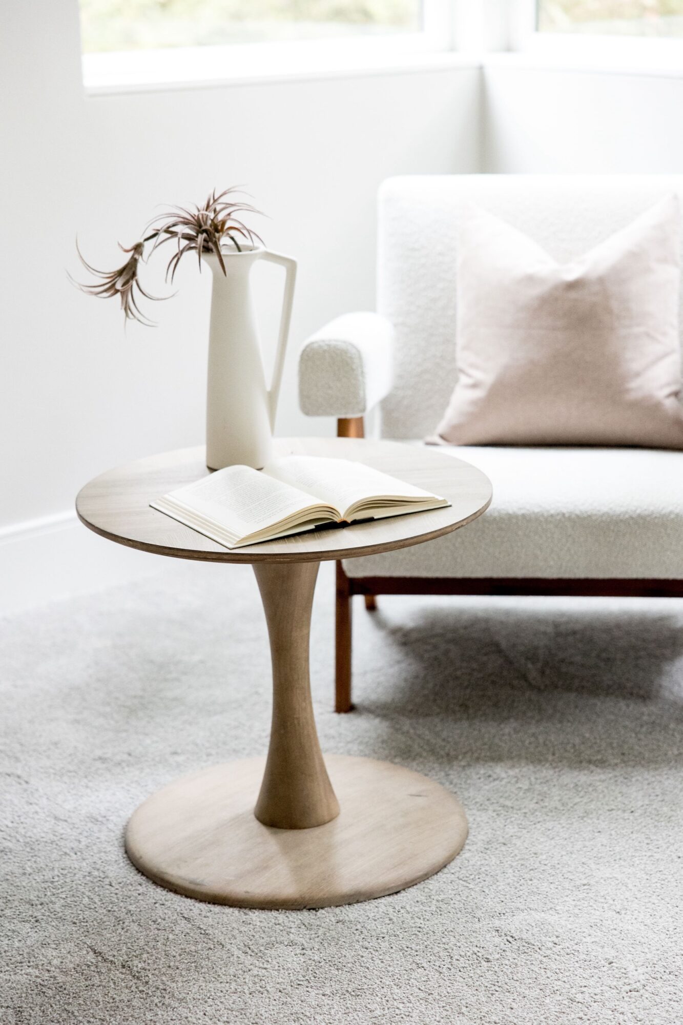 Seattle's best commercial food and hospitality photographer A round wooden side table with an open book and a tall white vase holding dried foliage, styled by Seattles best commercial food lifestyle photographer, sits next to a white armchair with a light pink pillow on a light carpeted floor by a window. captured by seattle's best food and lifestyle photographer Brooke Fitts