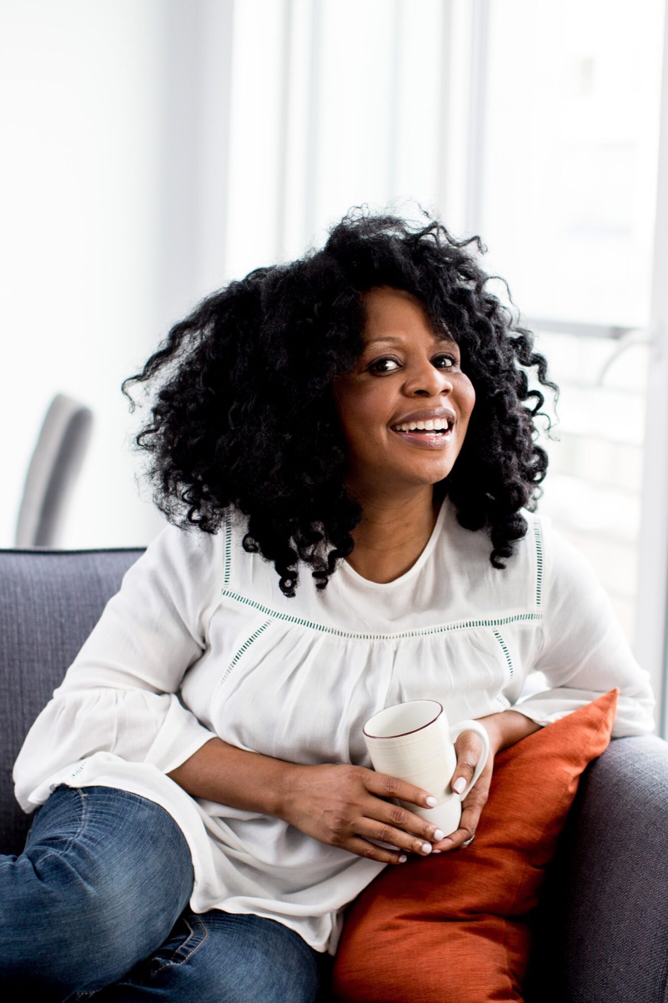 Seattle's best commercial food and hospitality photographer A woman with curly hair sits on a gray couch, smiling and holding a white mug, as natural light pours through large windows—captured by Seattles best commercial food lifestyle photographer. captured by seattle's best food and lifestyle photographer Brooke Fitts