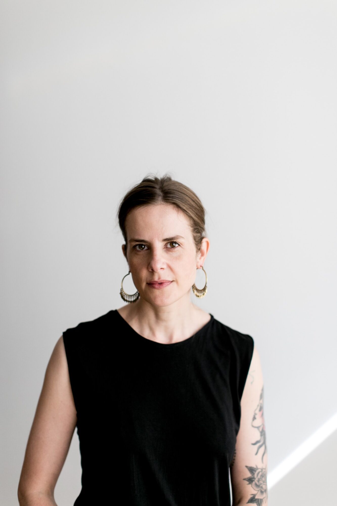 Seattle's best commercial food and hospitality photographer A woman with light skin, brown hair tied back, wearing large hoop earrings and a black sleeveless top, stands against a plain white background. Captured by Seattles best commercial food lifestyle photographer, a floral tattoo is visible on her left arm. captured by seattle's best food and lifestyle photographer Brooke Fitts