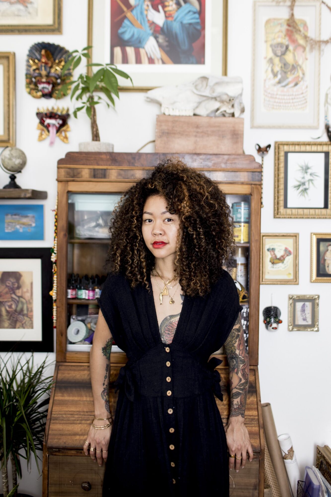 Seattle's best commercial food and hospitality photographer A woman with curly hair wearing a black dress stands in front of a wooden cabinet, surrounded by eclectic framed art and plants on a white wall—captured by Seattles best commercial food lifestyle photographer. captured by seattle's best food and lifestyle photographer Brooke Fitts