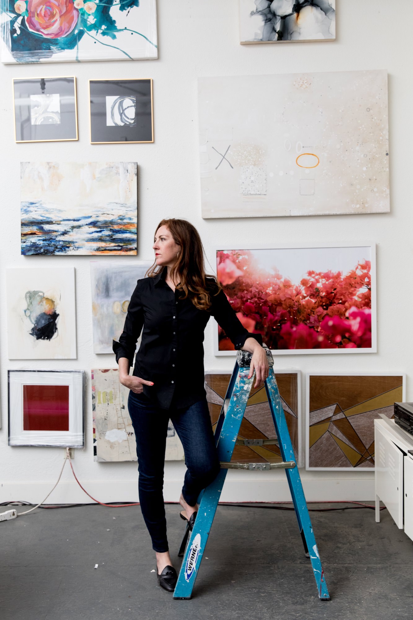 Seattle's best commercial food and hospitality photographer A woman in a black shirt and jeans stands on a blue step ladder in an art studio, surrounded by colorful abstract paintings—captured by Seattles best commercial food lifestyle photographer. captured by seattle's best food and lifestyle photographer Brooke Fitts