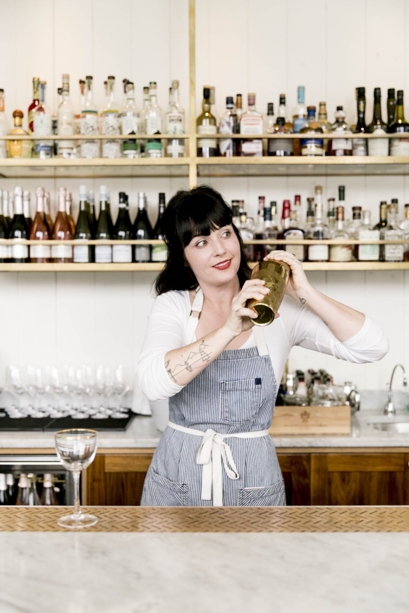Seattle's best commercial food and hospitality photographer A bartender with dark hair and bangs, wearing a striped apron over a white shirt, shakes a cocktail shaker behind a bar lined with bottles and glassware, captured by Seattles best commercial food lifestyle photographer. captured by seattle's best food and lifestyle photographer Brooke Fitts