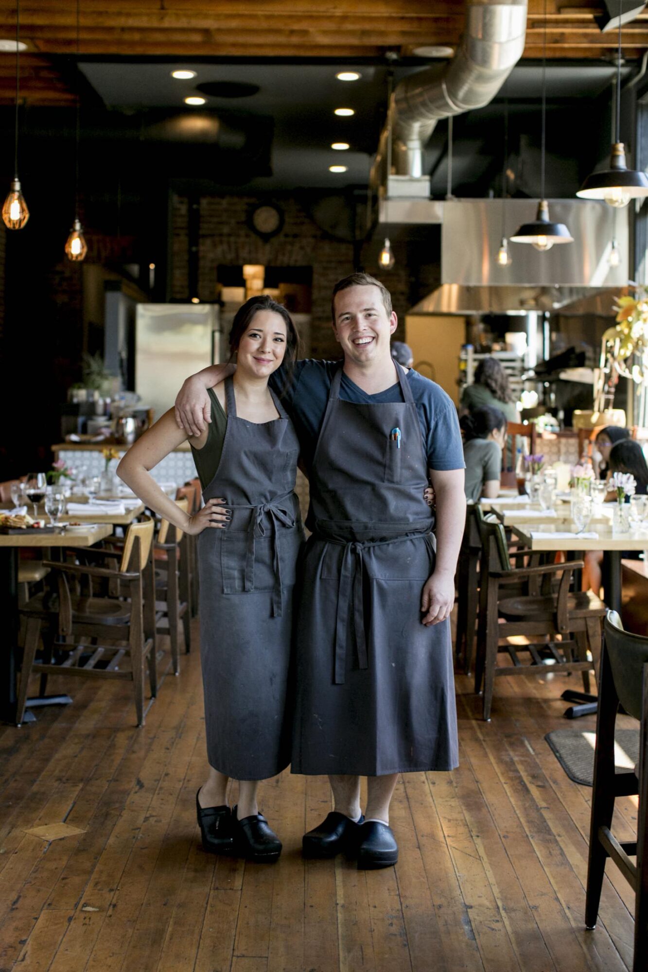 Seattle's best commercial food and hospitality photographer Two restaurant workers in dark aprons and clogs stand smiling with arms around each other in the middle of a cozy, rustic dining area—captured by Seattles best commercial food lifestyle photographer. captured by seattle's best food and lifestyle photographer Brooke Fitts