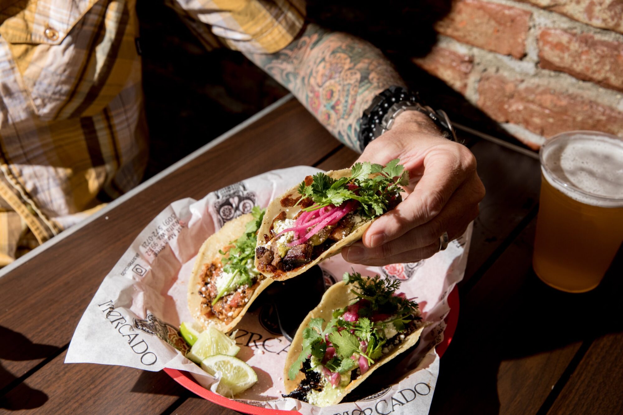 Seattle's best commercial food and hospitality photographer A person with a tattooed arm holds a taco filled with meat, onions, and cilantro over a basket of two more tacos and lime wedges, next to a pint of beer on a wooden table—captured by Seattles best commercial food lifestyle photographer. captured by seattle's best food and lifestyle photographer Brooke Fitts