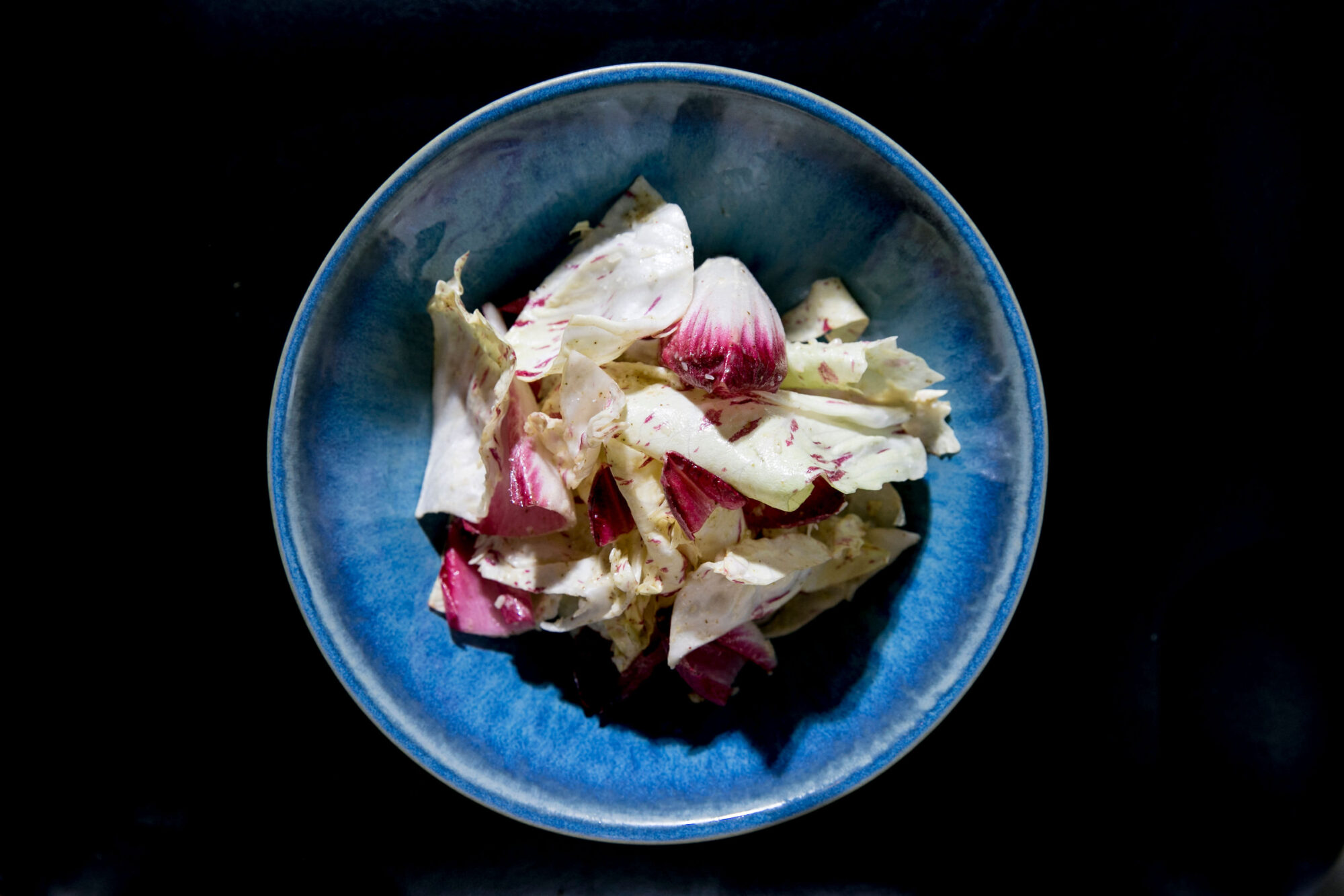 Seattle's best commercial food and hospitality photographer A blue bowl filled with light green and purple radicchio leaves sits against a dark background, illuminated by a direct overhead light—captured by Seattles best commercial food lifestyle photographer. captured by seattle's best food and lifestyle photographer Brooke Fitts