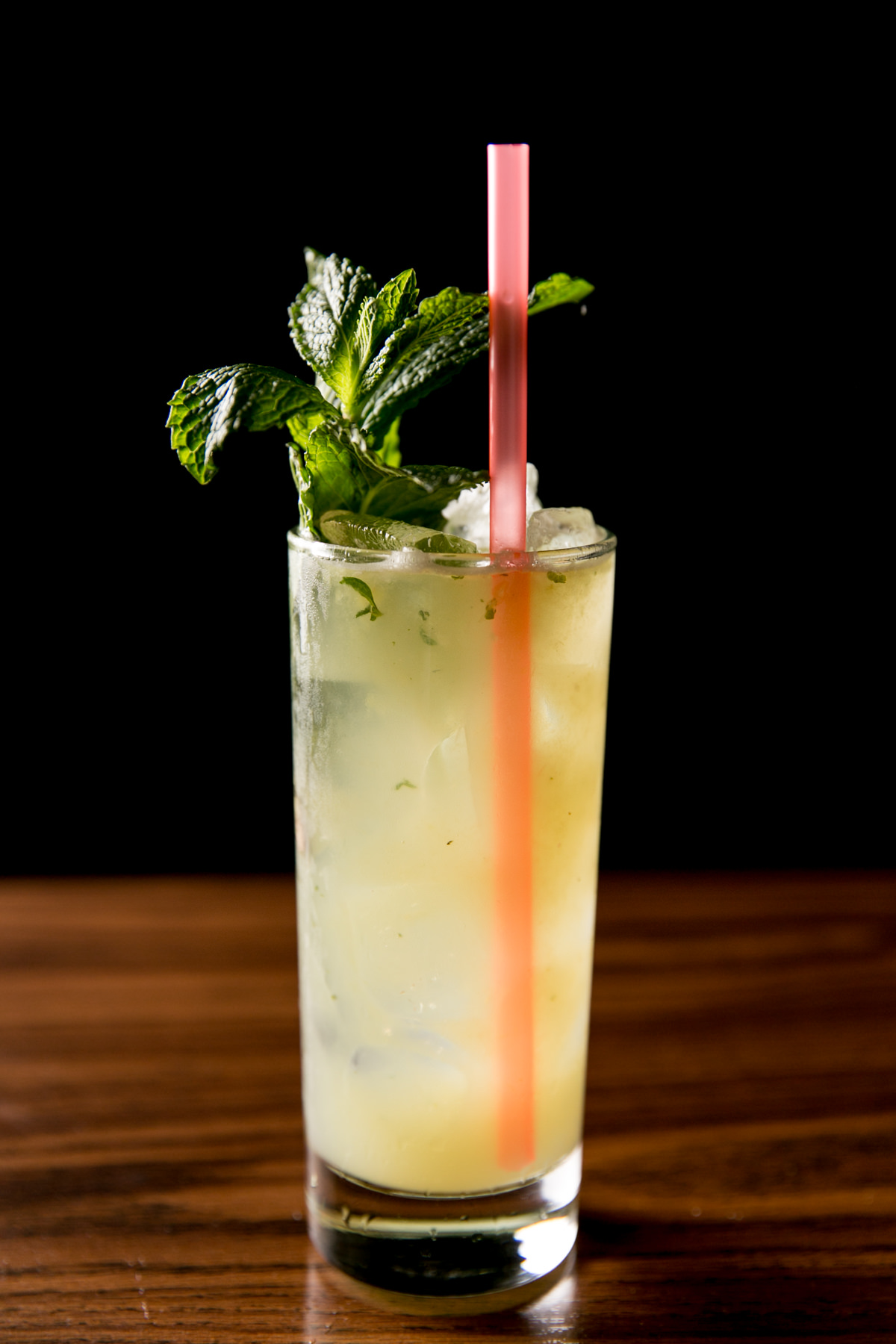 Seattle's best commercial food and hospitality photographer A tall glass of light yellow cocktail with crushed ice, fresh mint garnish, and a pink straw sits on a wooden surface against a black background—captured by Seattles best commercial food lifestyle photographer. captured by seattle's best food and lifestyle photographer Brooke Fitts