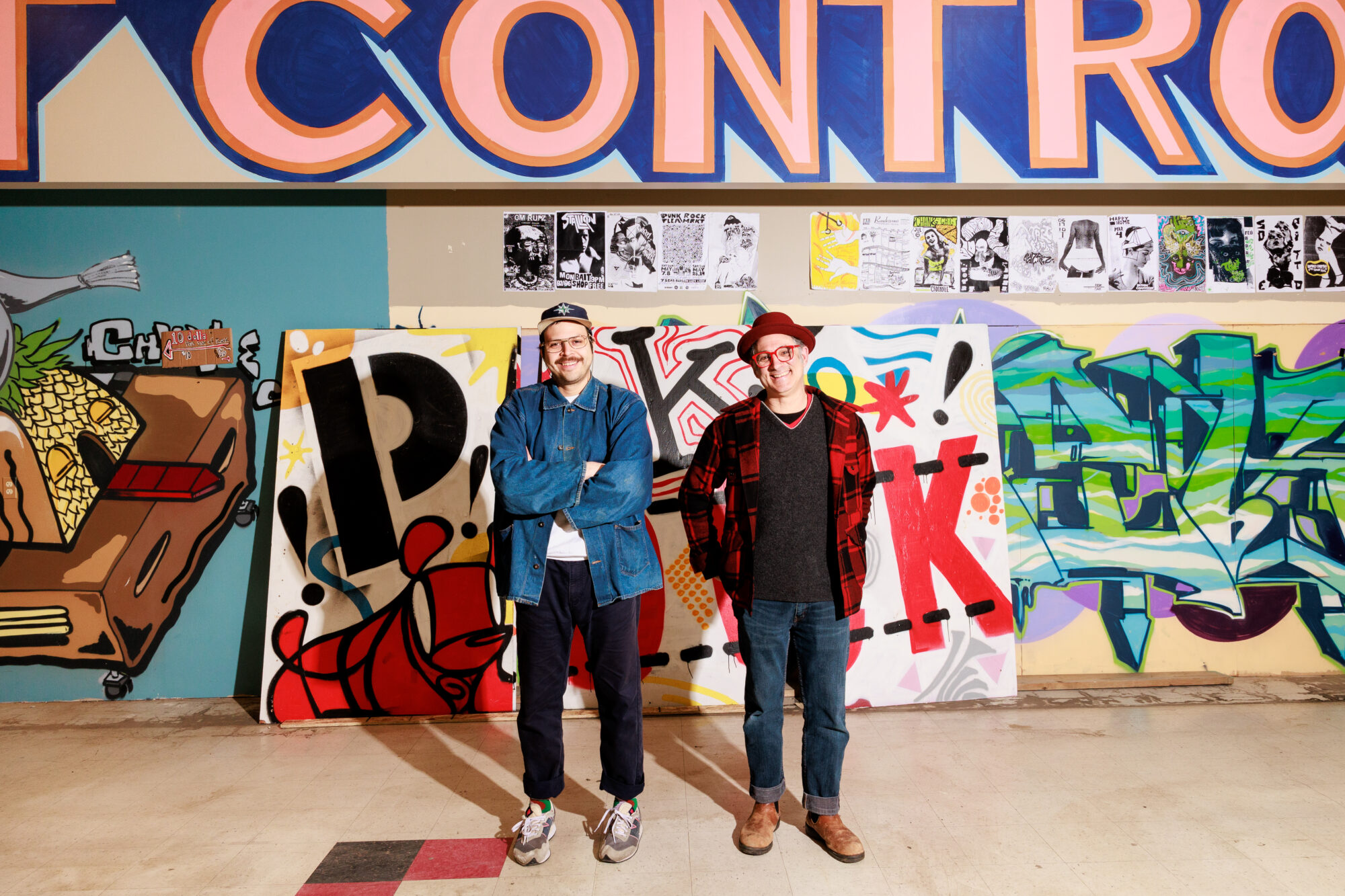 Seattle's best commercial food and hospitality photographer Two men stand side by side, smiling in front of a colorful graffiti wall with bold artwork and posters. Captured by Seattles best commercial food lifestyle photographer, a large sign above them reads CONTROL in partial view. captured by seattle's best food and lifestyle photographer Brooke Fitts