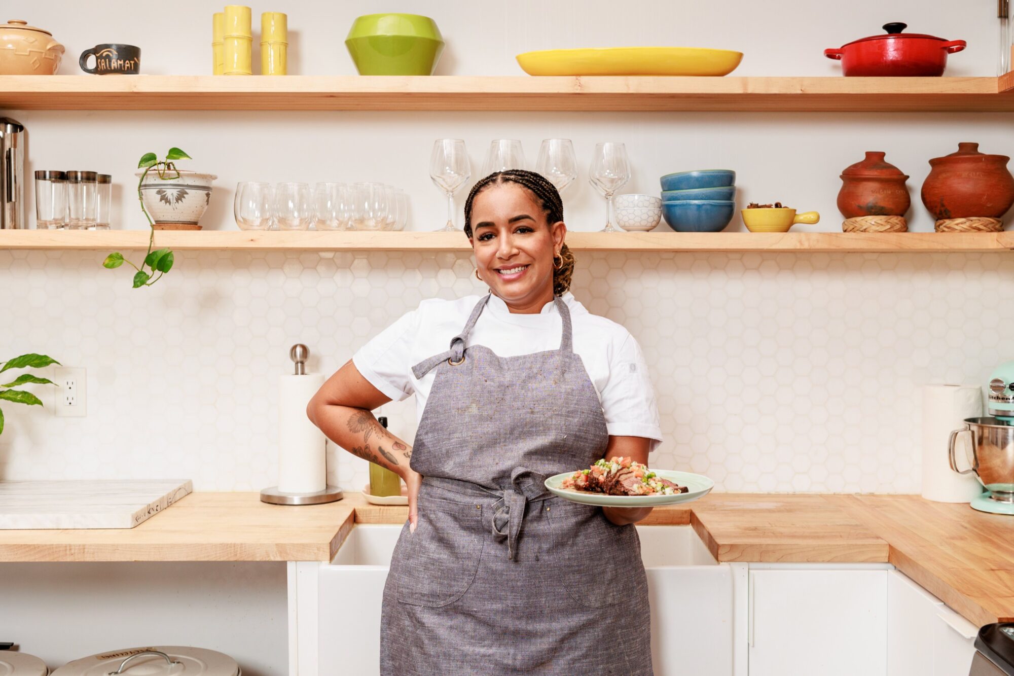 Seattle's best commercial food and hospitality photographer A smiling woman in a gray apron stands in a modern kitchen, holding a plate of food. Captured by Seattles best commercial food lifestyle photographer, shelves behind her display dishes and cups on light wood counters with a white hexagonal tile backsplash. captured by seattle's best food and lifestyle photographer Brooke Fitts