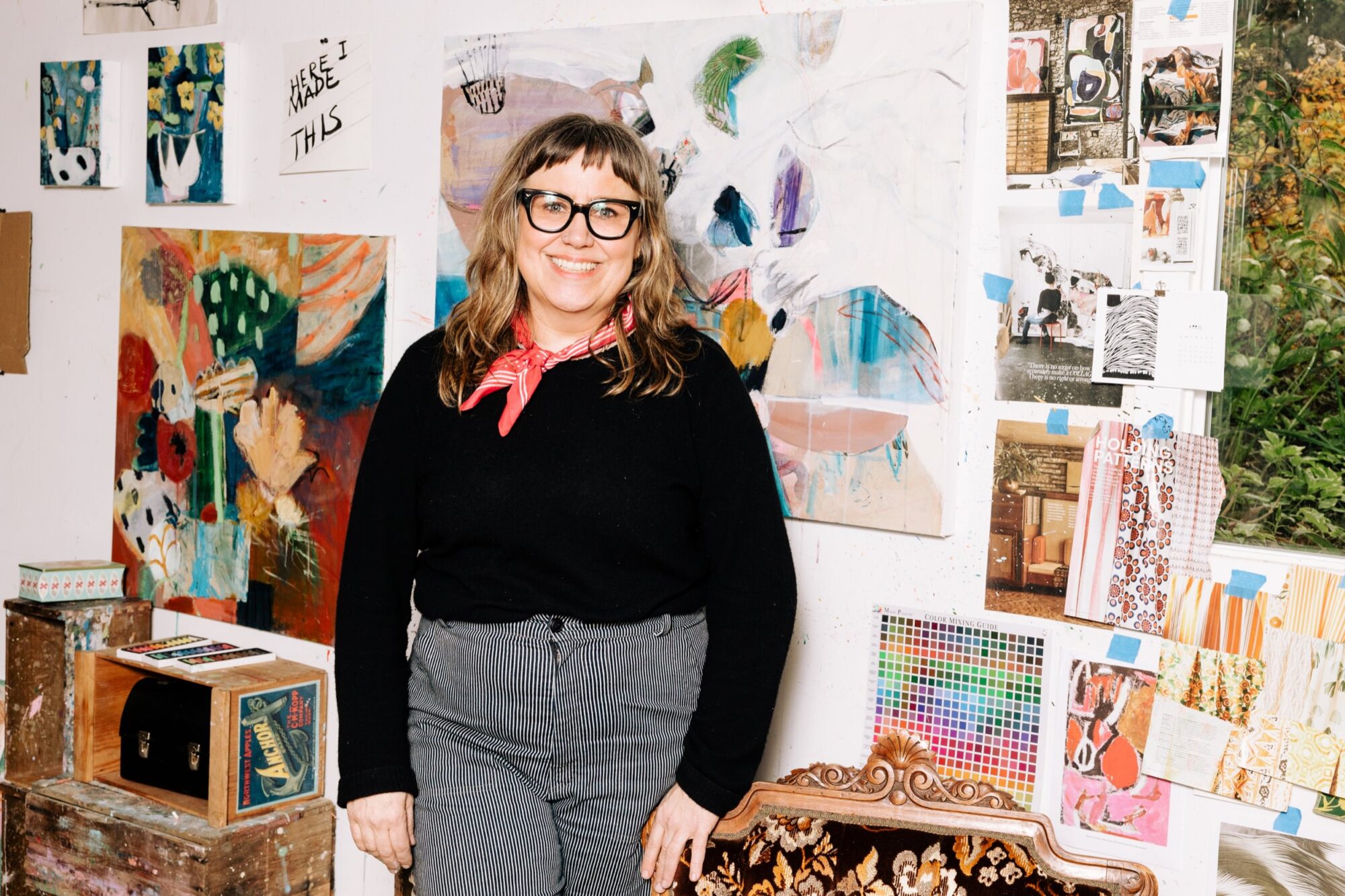 Seattle's best commercial food and hospitality photographer A woman with glasses, a red scarf, and striped pants smiles while standing in an art studio filled with colorful paintings and inspiration photos—captured by Seattles best commercial food lifestyle photographer. captured by seattle's best food and lifestyle photographer Brooke Fitts