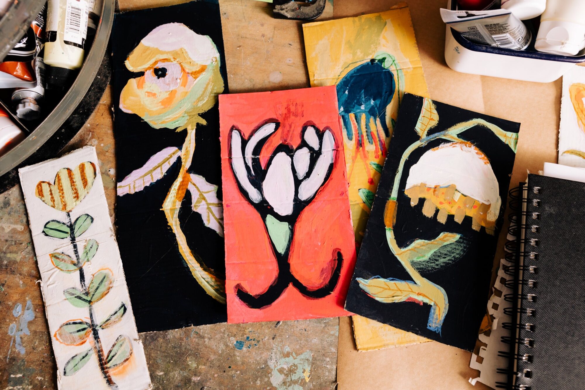 Seattle's best commercial food and hospitality photographer A tabletop with colorful abstract floral paintings on small canvases, surrounded by art supplies, a sketchbook, and paint tubes—styled as if curated by Seattles best commercial food lifestyle photographer. The artworks feature bold brushstrokes and vibrant colors. captured by seattle's best food and lifestyle photographer Brooke Fitts