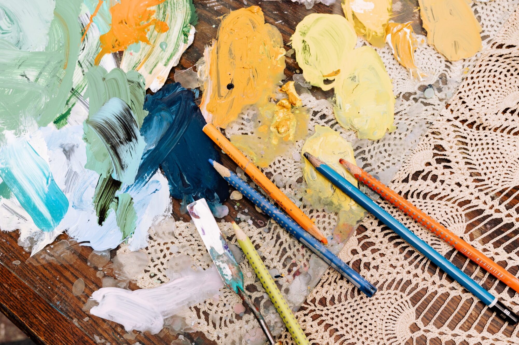Seattle's best commercial food and hospitality photographer Colored pencils and a palette knife rest on a wooden table covered with paint smudges in green, yellow, blue, and white, styled by Seattles best commercial food lifestyle photographer, alongside a cream-colored crocheted lace doily. captured by seattle's best food and lifestyle photographer Brooke Fitts