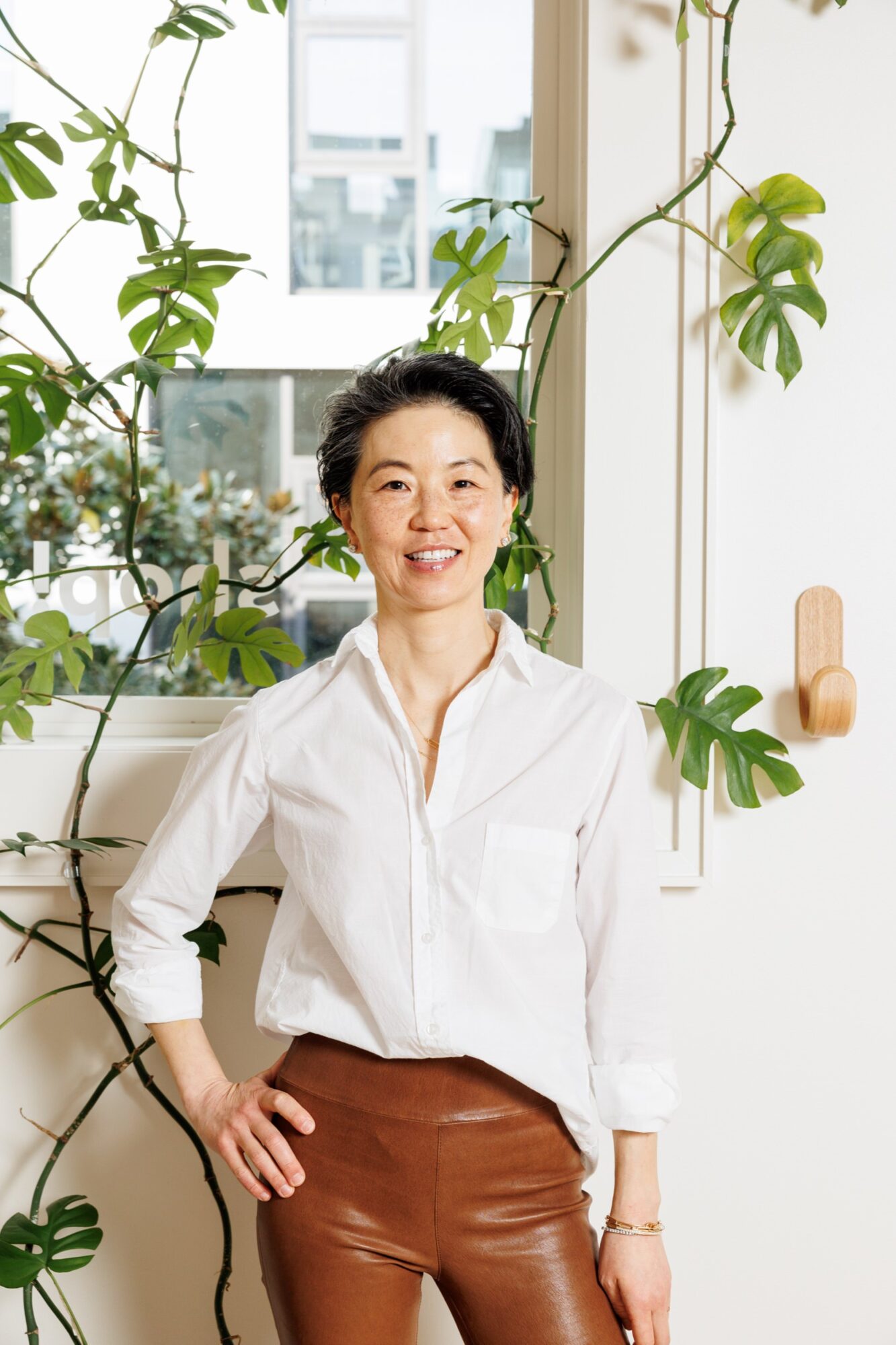 Seattle's best commercial food and hospitality photographer A woman with short dark hair stands indoors, smiling. She is wearing a white button-up shirt and brown pants. Behind her are large green leafy plants and a bright window, captured by Seattles best commercial food lifestyle photographer. captured by seattle's best food and lifestyle photographer Brooke Fitts