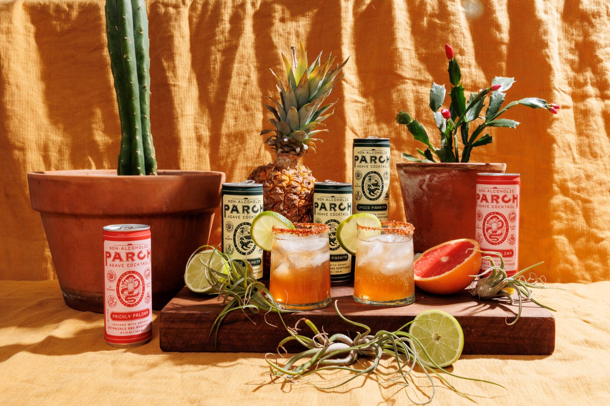 Seattle's best commercial food and hospitality photographer A colorful tabletop display by Seattles best commercial food lifestyle photographer features cans of Parch Prickly Paloma, cocktail glasses garnished with lime, sliced grapefruit, pineapples, potted cacti, and scattered limes on a yellow fabric backdrop. captured by seattle's best food and lifestyle photographer Brooke Fitts