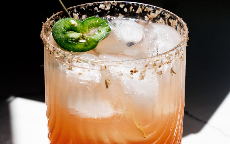Seattle's best commercial food and hospitality photographer A glass of iced cocktail with a salted rim, orange-pink gradient, garnished with jalapeño slices and a decorative metal stirrer, is beautifully captured in dramatic light by Seattles best commercial food lifestyle photographer. captured by seattle's best food and lifestyle photographer Brooke Fitts