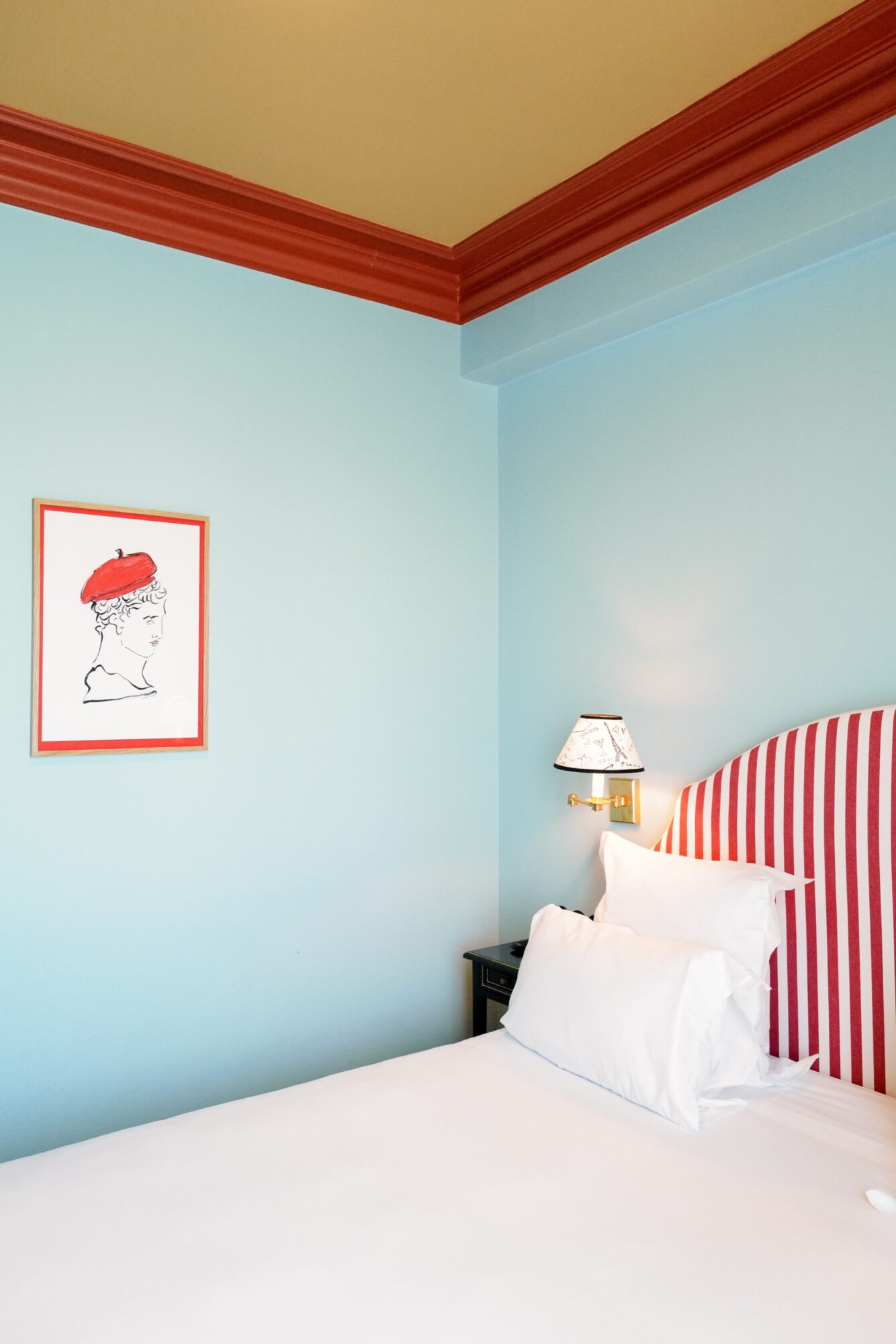 Seattle's best commercial food and hospitality photographer A neatly made bed with white linens and pillows, a red and white striped headboard, a small wall-mounted lamp, and a framed sketch of a person in a red hat—captured by Seattles best commercial food lifestyle photographer—against a pale blue wall. captured by seattle's best food and lifestyle photographer Brooke Fitts