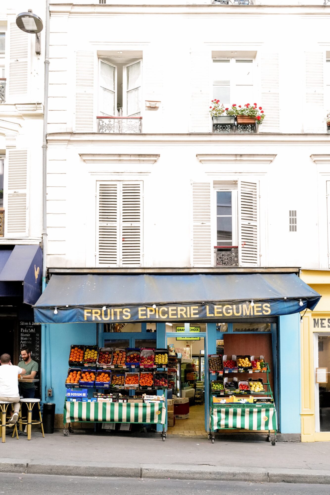 Seattle's best commercial food and hospitality photographer A small Parisian fruit and vegetable shop with a blue awning reading FRUITS ÉPICERIE LÉGUMES is pictured as if by Seattles best commercial food lifestyle photographer—colorful produce outside, white shutters, and flower boxes above. captured by seattle's best food and lifestyle photographer Brooke Fitts
