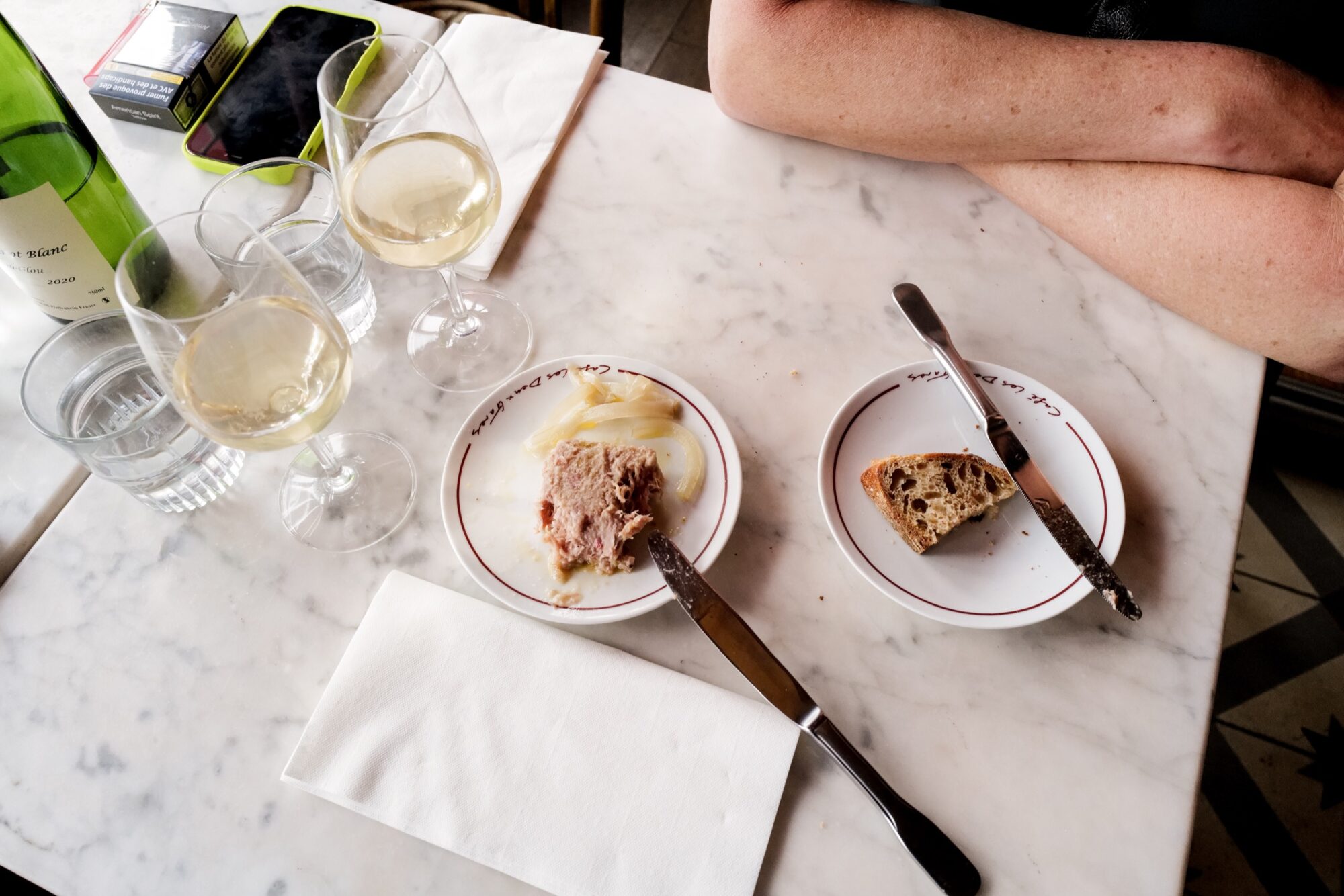 Seattle's best commercial food and hospitality photographer A marble table with three glasses of white wine, a bottle, plates of pâté and bread, cutlery, a glass of water, and a napkin—captured by Seattles best commercial food lifestyle photographer as someone rests their crossed arms nearby. captured by seattle's best food and lifestyle photographer Brooke Fitts