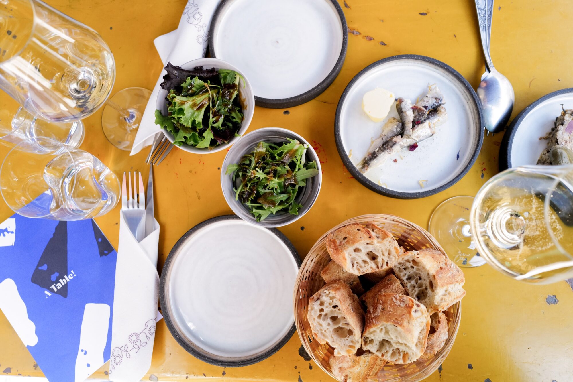Seattle's best commercial food and hospitality photographer A yellow table set with empty white plates, a basket of sliced bread, two bowls of green salad, a plate with sardines and butter, drinking glasses, utensils, blue-and-white napkins—captured by Seattles best commercial food lifestyle photographer. captured by seattle's best food and lifestyle photographer Brooke Fitts