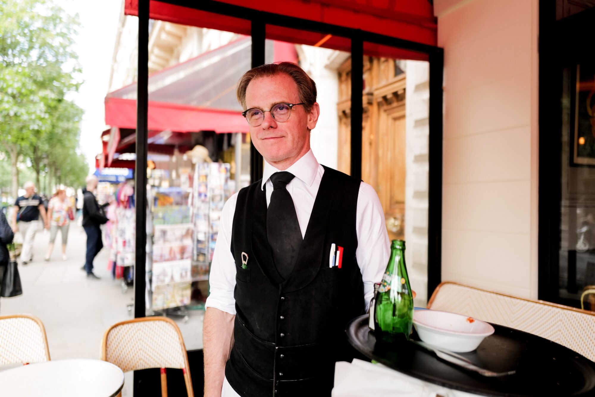 Seattle's best commercial food and hospitality photographer A waiter in a black vest and white shirt stands at an outdoor café table with a tray holding a green bottle and bowl, captured by Seattles best commercial food lifestyle photographer. Behind him, a vibrant city street bustles with people and café seating. captured by seattle's best food and lifestyle photographer Brooke Fitts