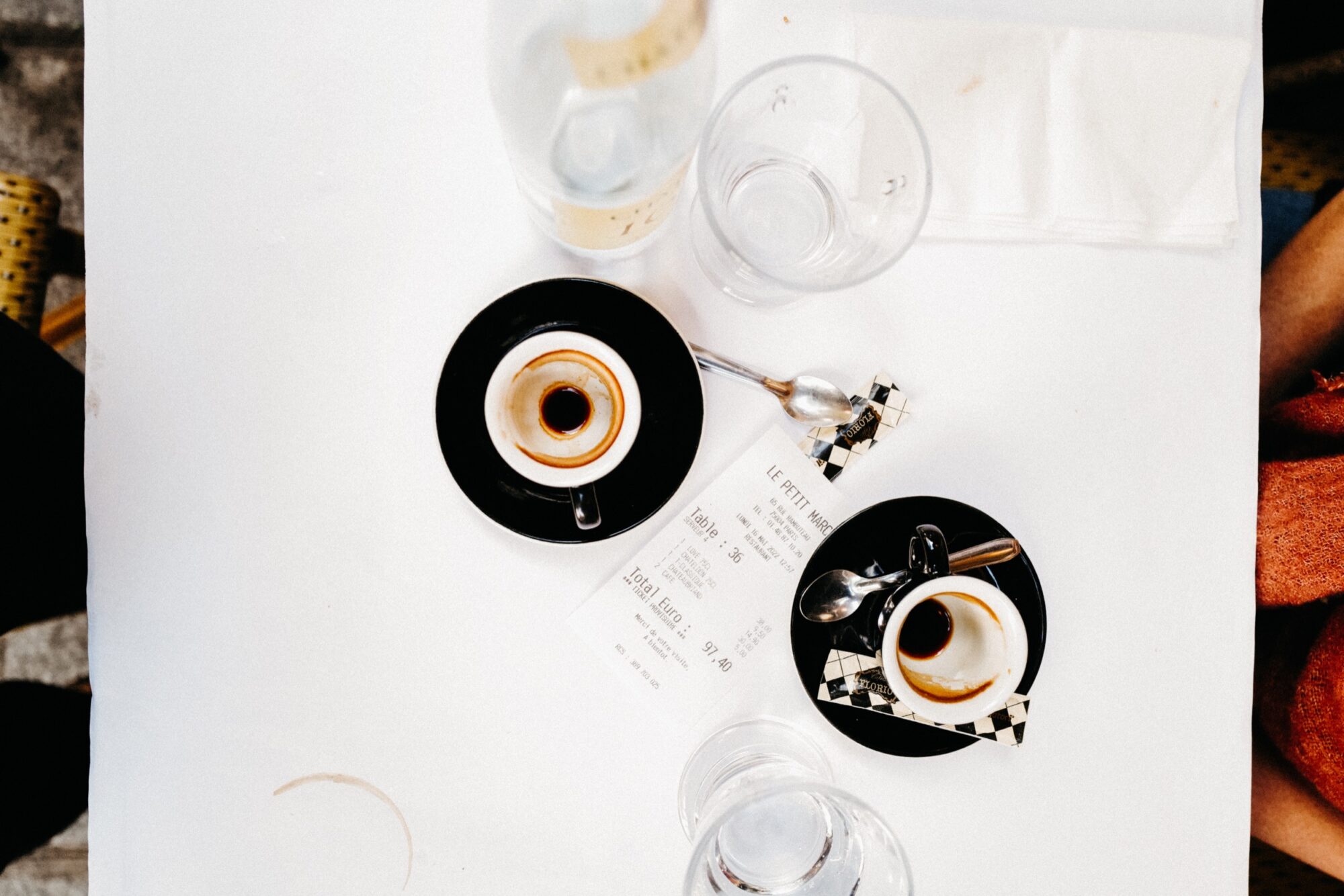Seattle's best commercial food and hospitality photographer A top-down view of a white table styled by Seattles best commercial food lifestyle photographer, featuring two empty espresso cups on black saucers, glasses of water, a receipt, napkins, and a spoon. Two people are partly visible at the table’s edge. captured by seattle's best food and lifestyle photographer Brooke Fitts