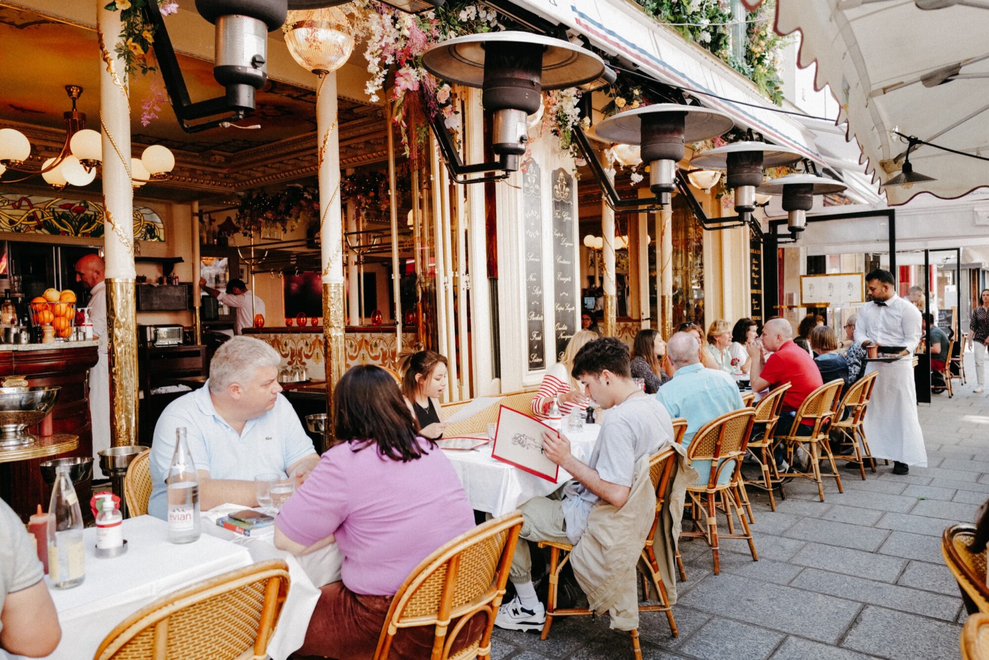 Seattle's best commercial food and hospitality photographer People sit at outdoor tables of a lively, colorful sidewalk café—some reading menus, others chatting—as a waiter in a white apron approaches with a tray. Captured by Seattles best commercial food lifestyle photographer, flowers and ornate details set the scene. captured by seattle's best food and lifestyle photographer Brooke Fitts