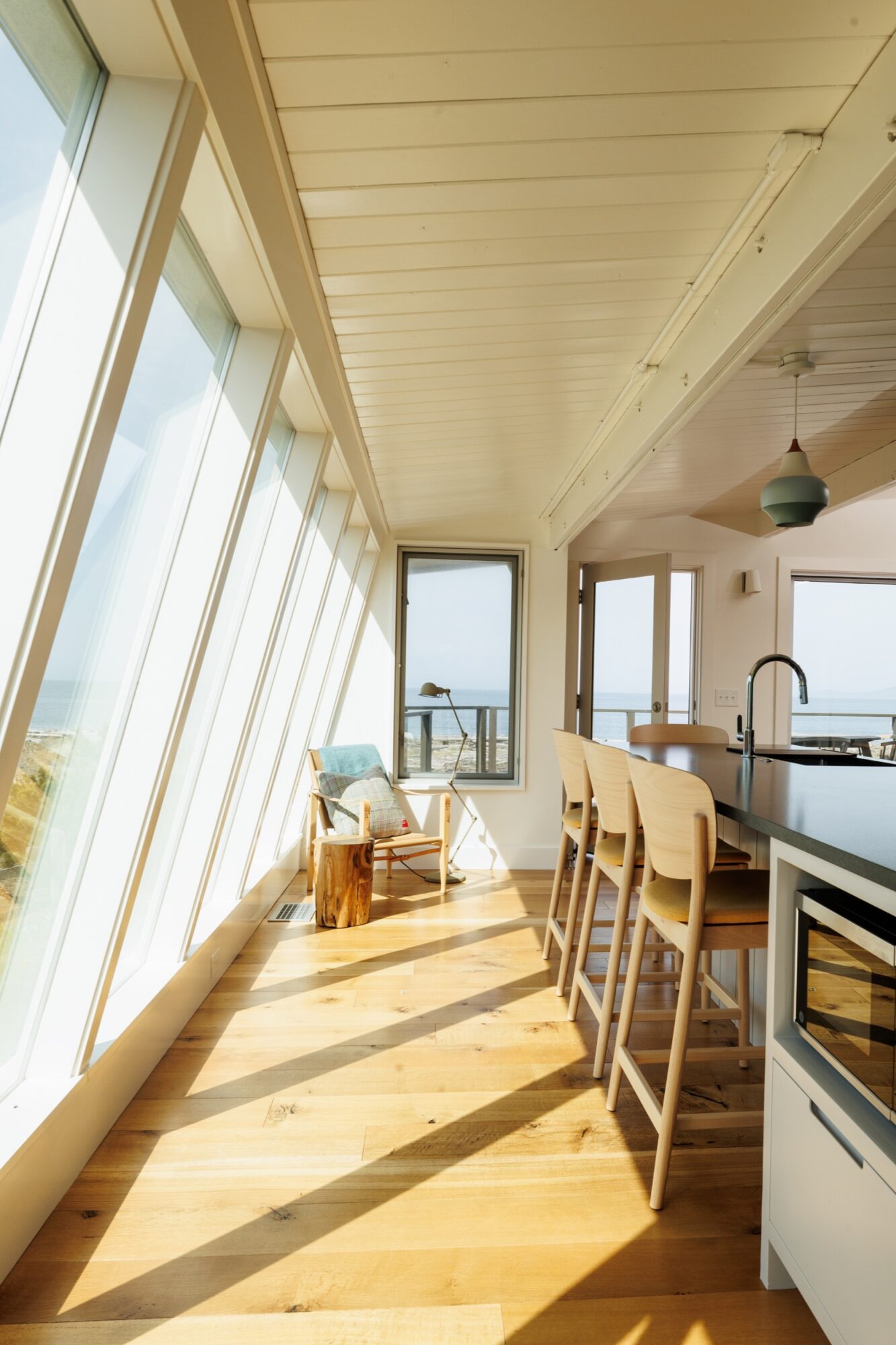 Seattle's best commercial food and hospitality photographer A bright, modern kitchen and dining area with large slanted windows, wooden floors, and high chairs at a counter, captured by a Seattle commercial interior design photographer for restaurants and hotels, all overlooking a scenic ocean view. captured by seattle's best food and lifestyle photographer Brooke Fitts