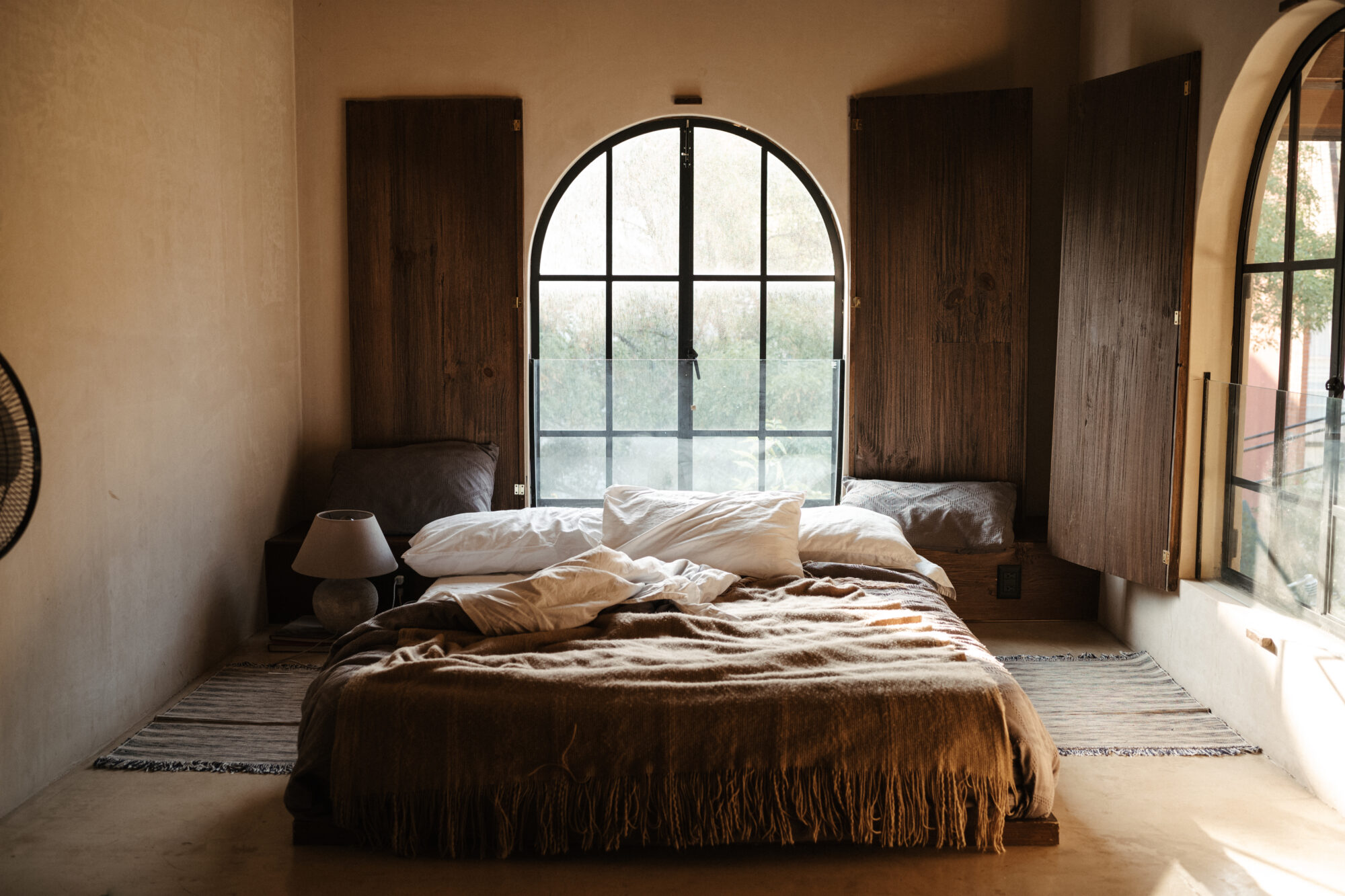 Seattle's best commercial food and hospitality photographer Cozy bedroom with an unmade bed, brown blanket, two pillows, and a bedside lamp. Sunlight streams through a large arched window with wooden shutters—capturing the rustic, minimalist vibe like Seattles best commercial food lifestyle photographer. captured by seattle's best food and lifestyle photographer Brooke Fitts