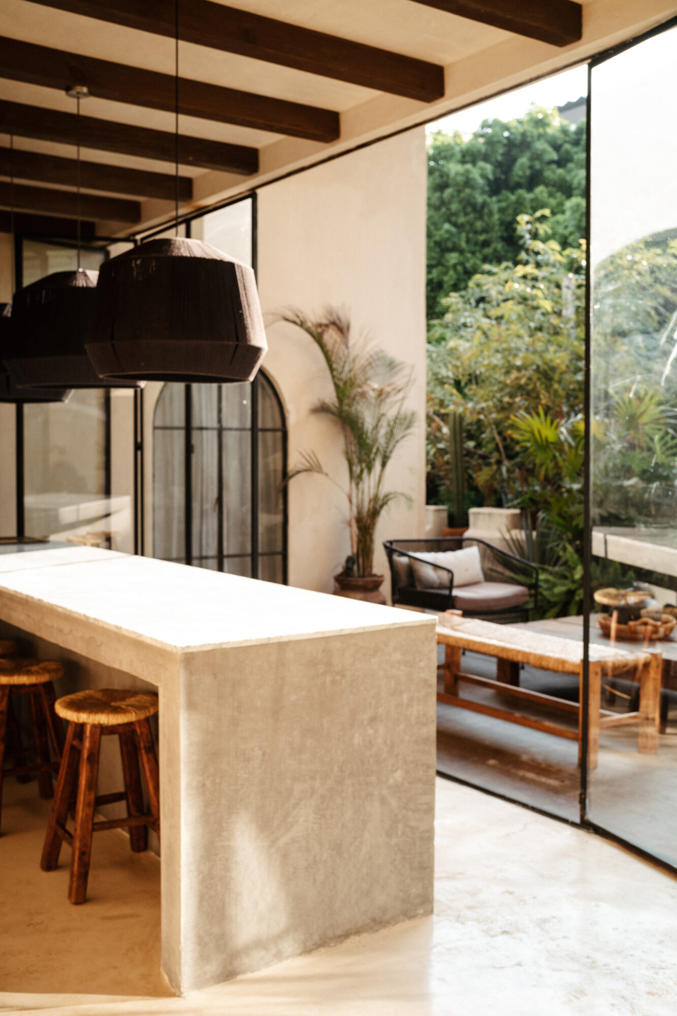 Seattle's best commercial food and hospitality photographer Modern kitchen with a concrete island and wooden stools, captured by Seattles best commercial food lifestyle photographer, features large black pendant lights and glass walls opening to a lush patio with wooden furniture and green plants. captured by seattle's best food and lifestyle photographer Brooke Fitts